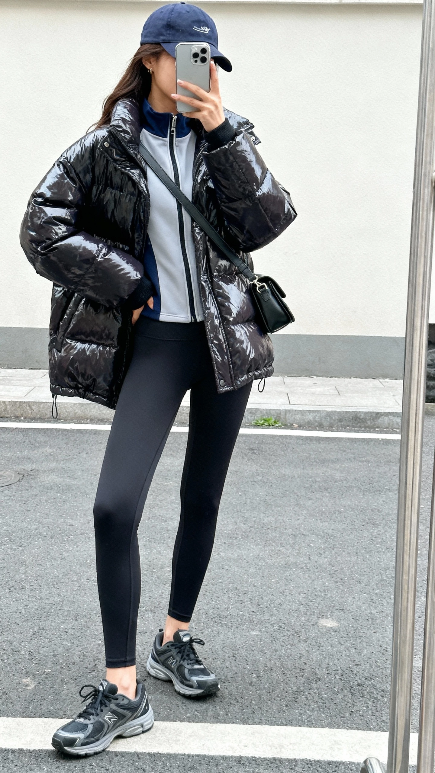 iPhone street-style side-angle selfie of a woman in a glossy puffer jacket layered over a sporty zip-up, leggings, sleek sneakers, baseball cap, and crossbody bag, face not visible, natural daylight, casual iPhone photo.