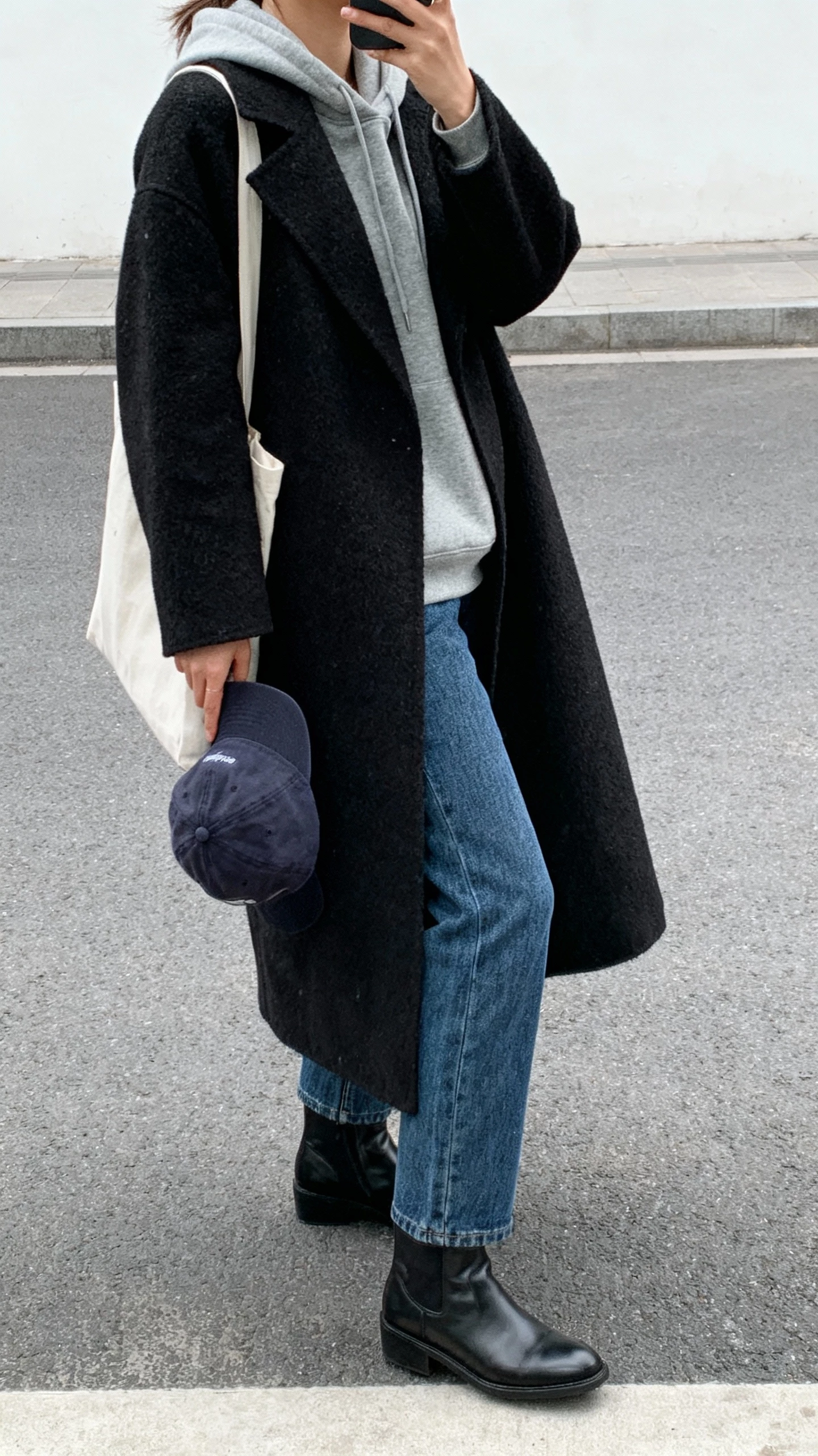 iPhone street-style side view selfie (phone held low) of a woman in a gray hoodie layered under a long black wool coat, straight blue jeans, clean black leather ankle boots, baseball cap in hand, minimal tote, face not visible, overcast daylight, natural iPhone photo.