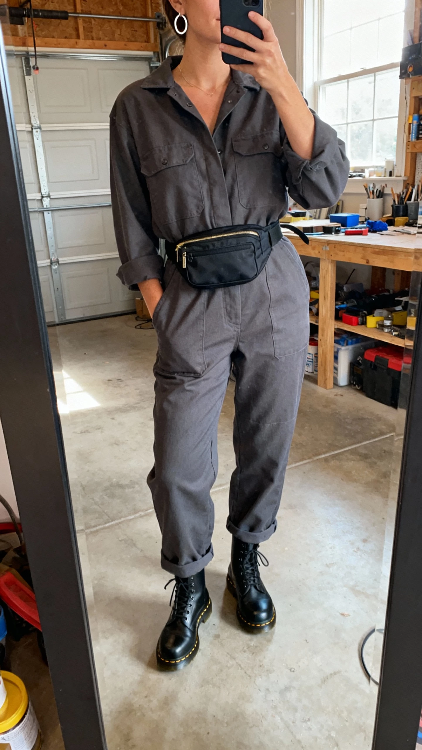 iPhone waist-up mirror selfie of a woman wearing a charcoal workwear jumpsuit cinched with a belt bag, black Doc Martens, simple hoop earrings, face not visible, taken in a garage/workspace with natural window light, natural iPhone photo quality.