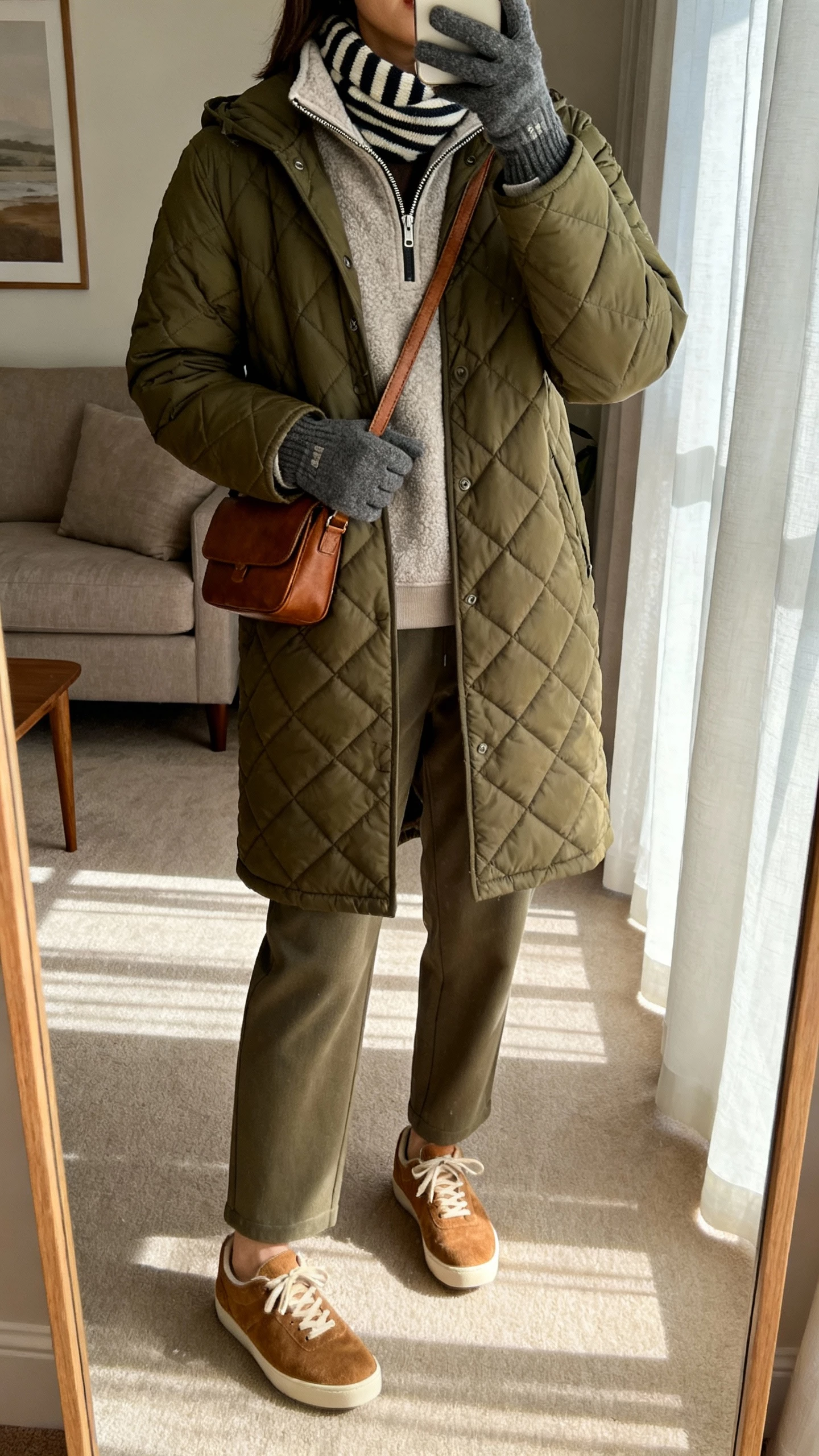 Living room mirror selfie of a woman wearing an olive quilted car coat, half-zip fleece, relaxed trousers, suede sneakers, leather crossbody, tech-friendly gloves, striped scarf, face not visible, neck-down crop, soft afternoon window light, natural iPhone photo.