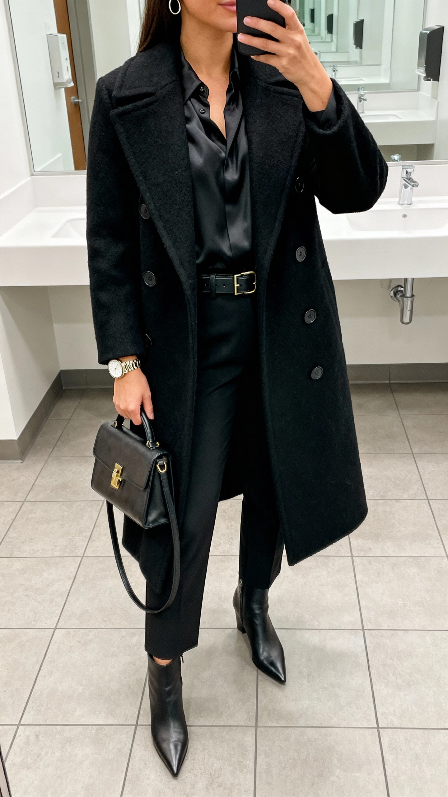 Office bathroom mirror selfie of a woman in a black double-breasted wool coat over a silk blouse, cigarette pants, pointed-toe ankle boots, leather belt, structured satchel, statement watch and small hoops, face not visible, neck-down crop, bright office lighting, natural iPhone photo.