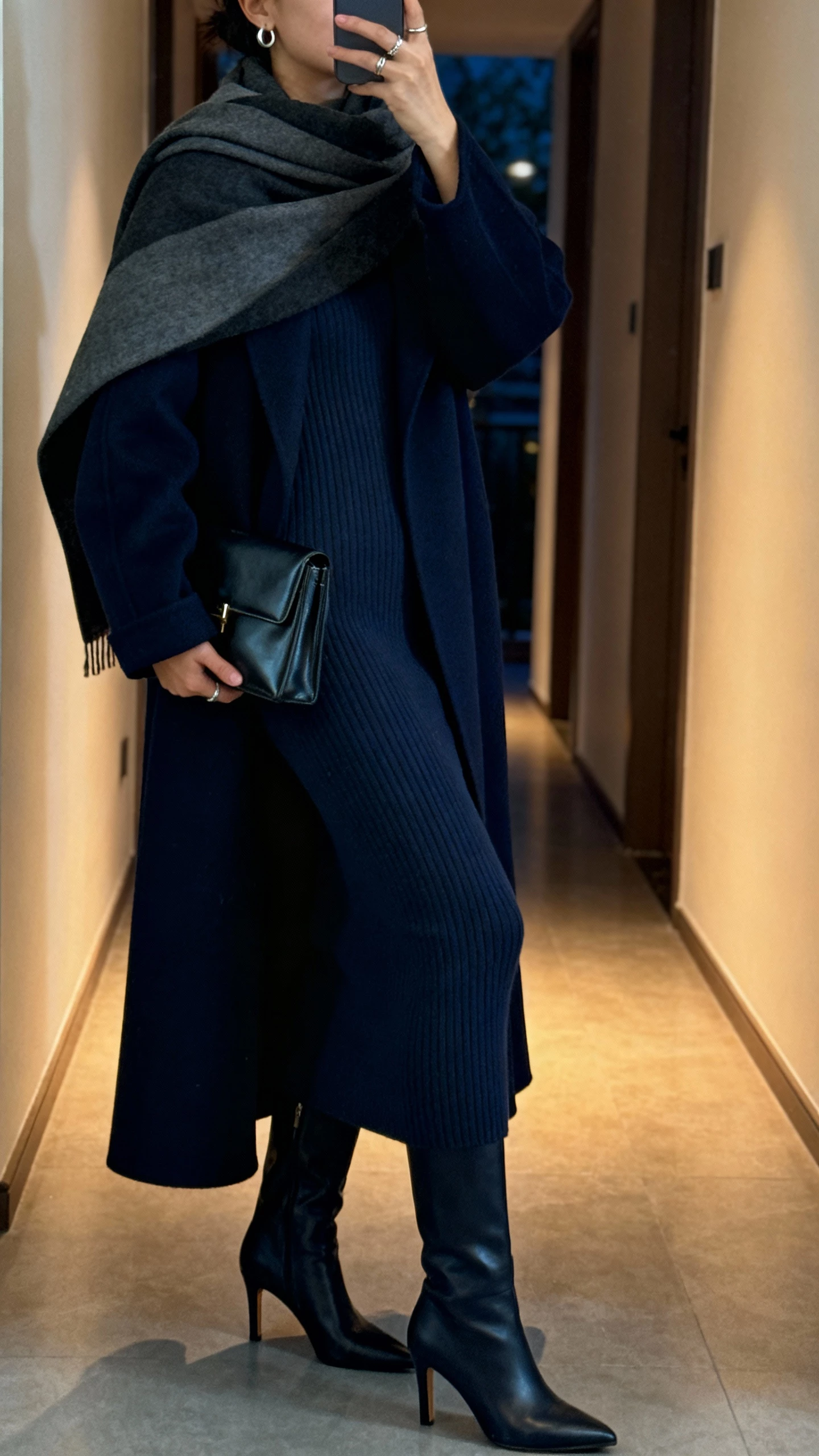 Side-angle corridor selfie of a woman wearing a deep navy maxi-length coat over a body-skimming knit dress, heeled knee boots, sleek clutch, minimalist ear cuffs and rings, optional wide scarf draped like a shawl, face not visible, evening hallway light, natural iPhone photo.