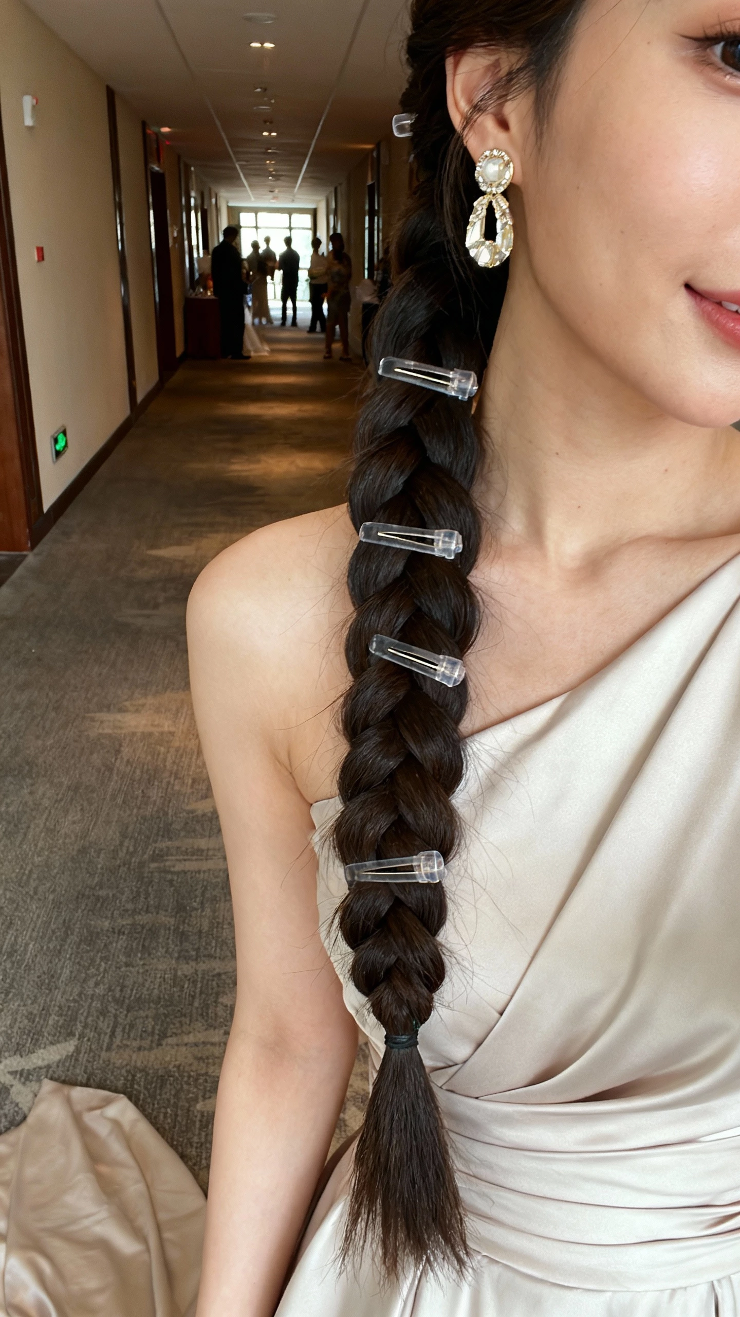 Side-angle iPhone selfie of a woman in a one-shoulder gown, long fishtail side braid over the shoulder with discreet clear elastics and hair pins, statement earrings, face not visible, venue corridor, natural indoor lighting, casual iPhone shot.