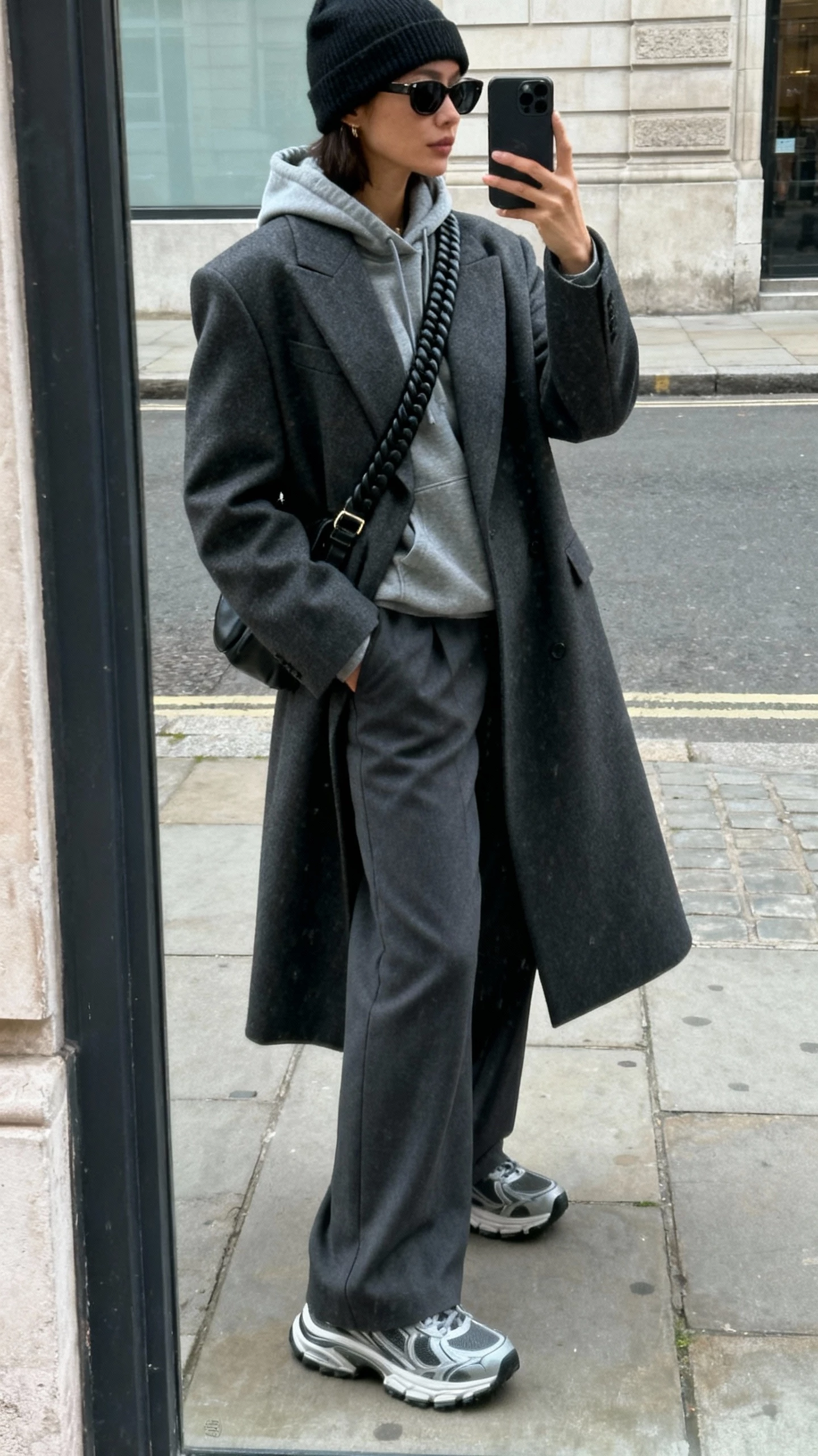 Street mirror-window selfie of a woman in an oversized tailored charcoal coat over a hoodie, wide-leg trousers, chunky sneakers, crossbody bag with thick strap, beanie and sunglasses, face not visible, side angle, downtown sidewalk, overcast daylight, natural iPhone photo.