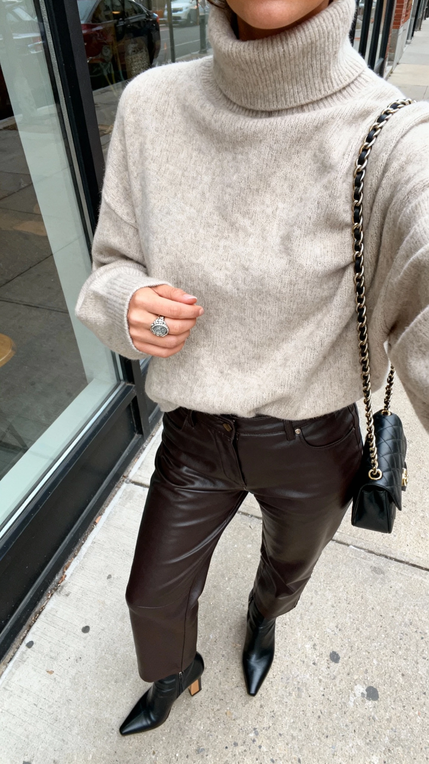 Street-style iPhone selfie cropped at shoulders showing a woman in an oversized turtleneck with high-waist leather pants, heeled ankle boots, chain-strap bag, statement ring, face not visible, city sidewalk window reflection, natural daylight, natural iPhone photo quality.