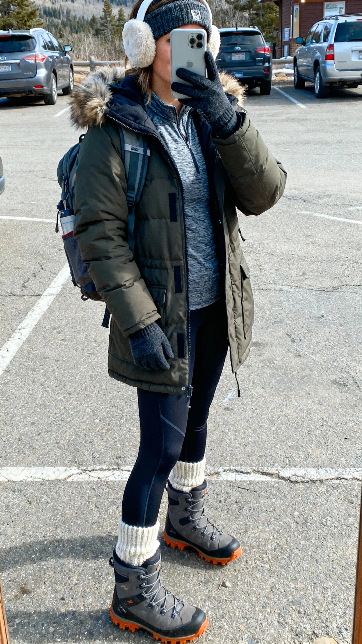 Trailhead parking lot selfie of a woman in an insulated parka with detachable hood trim, thermal base layer and cozy knit, fleece-lined leggings, weatherproof boots with good traction, backpack, ear warmer headband and touchscreen gloves, face not visible, outdoor daylight, natural iPhone quality.