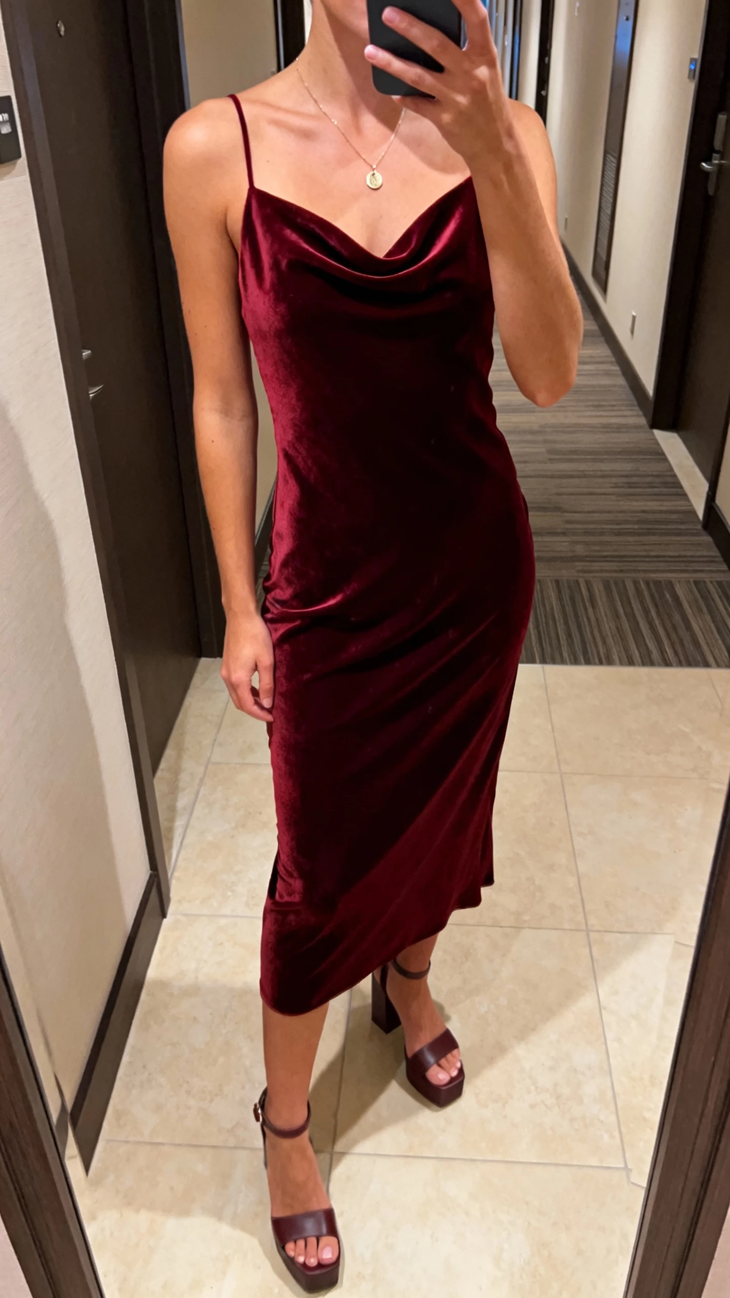 Casual iPhone selfie cropped at shoulders showing a woman in a burgundy velvet bias-cut slip dress with a subtle cowl, thin straps, simple pendant necklace, and block-heel sandals, face not visible, hotel hallway mirror, warm indoor lighting, natural iPhone aesthetic.