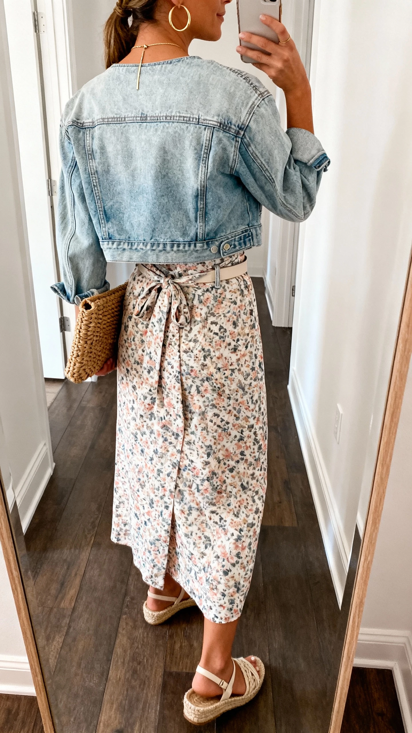 iPhone back-view hallway mirror selfie of a woman wearing a floral midi wrap dress with a cropped light-wash denim jacket, espadrille wedges, woven clutch, gold hoops, slim pendant necklace, optional lightweight belt, face not visible, natural daylight, casual iPhone photo.