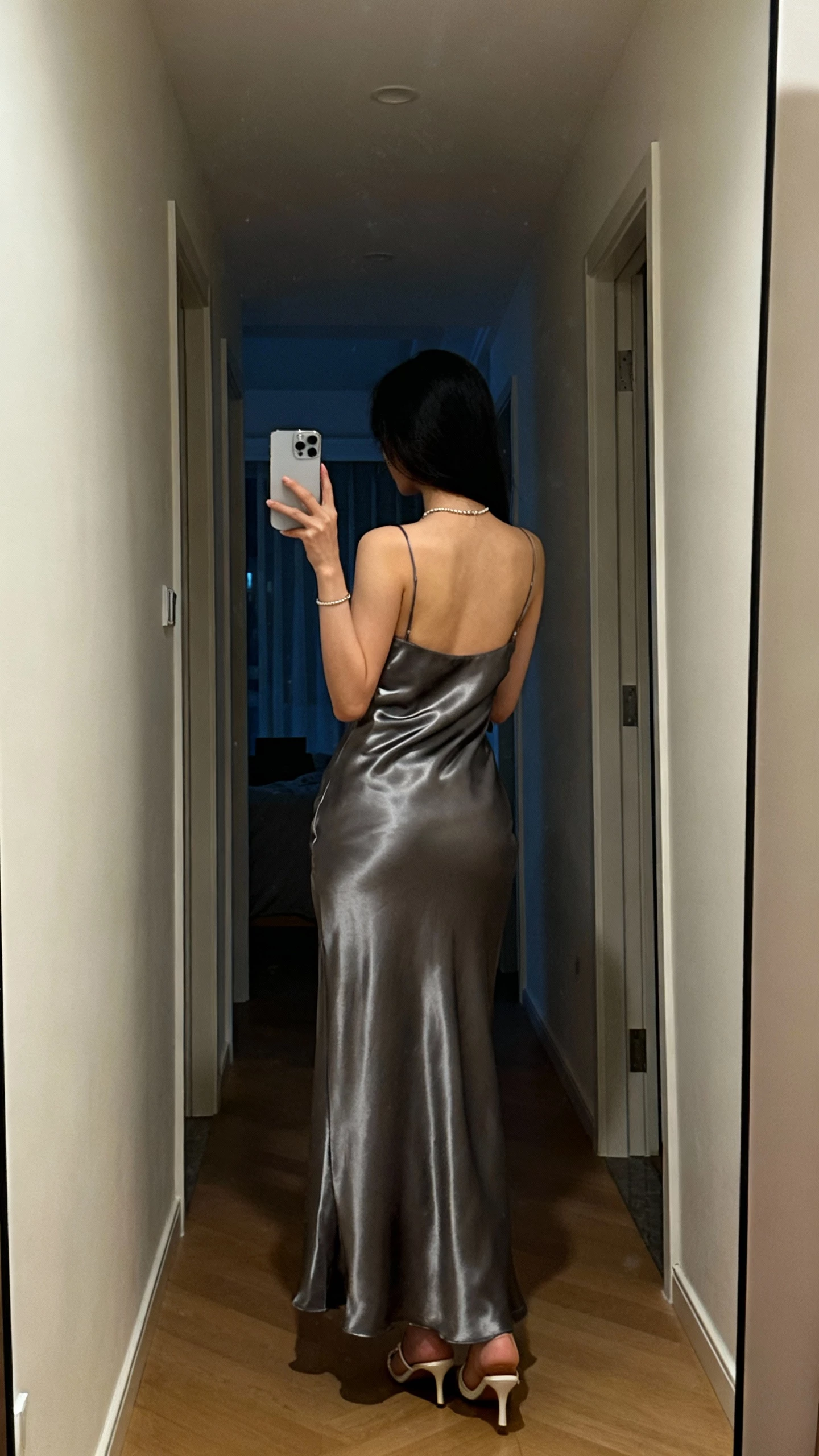 iPhone back-view hallway selfie of a woman in a gunmetal satin gown with minimal jewelry and simple heels, face not visible, soft evening indoor light, natural iPhone aesthetic