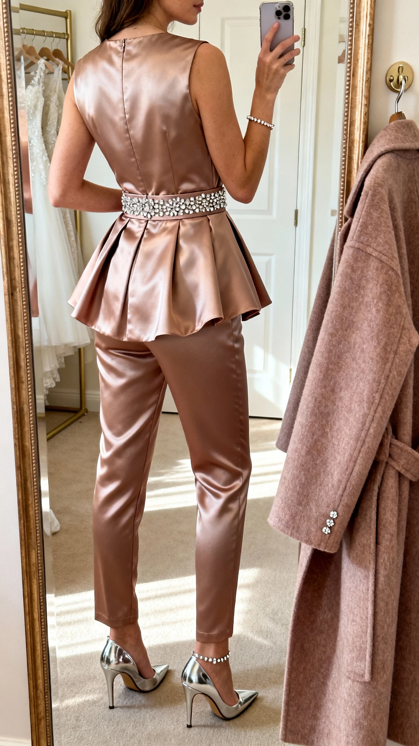 iPhone back-view mirror selfie of a woman in a structured satin jumpsuit with slim leg and detachable matching overskirt fastened at waist, embellished waistband, metallic classic stilettos, sparkling studs with delicate bracelet, long wool coat in coordinating tone on hook; face not visible, bridal suite mirror, soft morning light, natural iPhone photo quality.