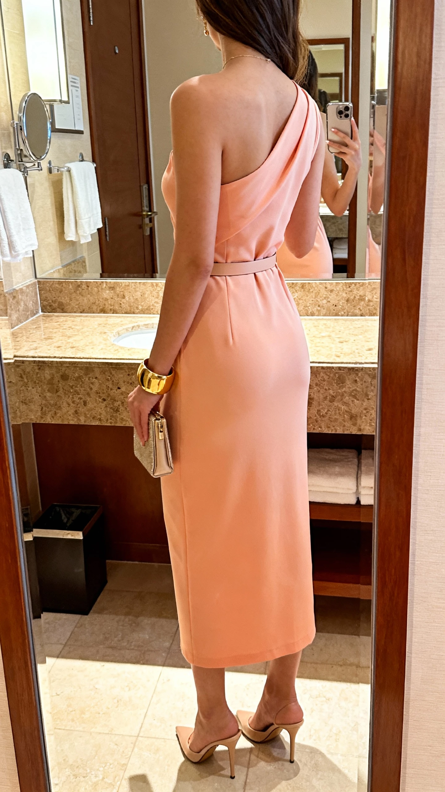 iPhone back-view mirror selfie of a woman wearing a soft peach one-shoulder column dress with minimal belt, gold cuff bracelet, sleek heels, small clutch, face not visible, hotel bathroom mirror with warm indoor light, casual iPhone shot.