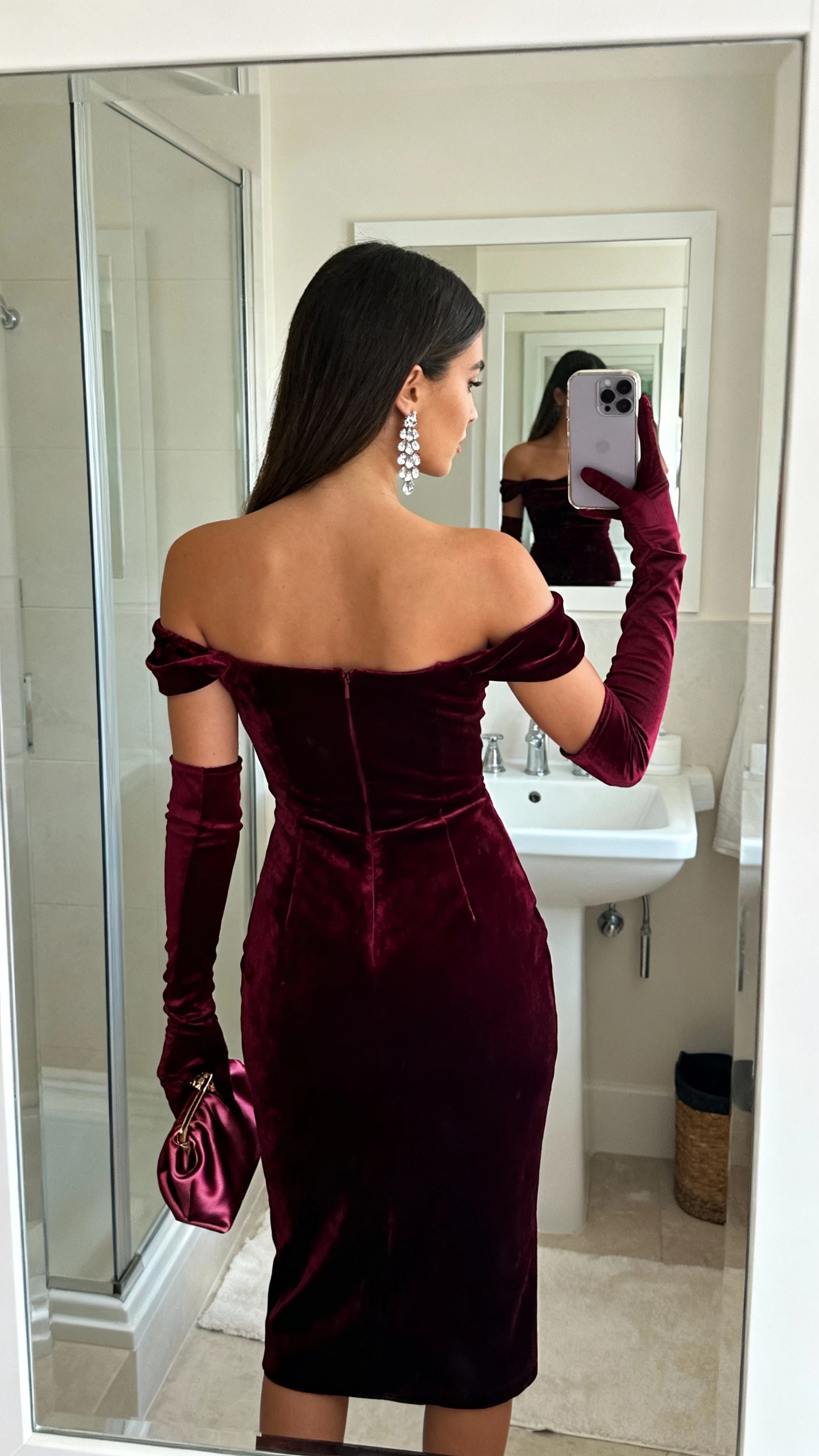 iPhone back-view selfie of a woman in a Bordeaux velvet off-the-shoulder Bardot midi with long opera gloves, crystal drop earrings, satin clutch, face not visible, bathroom mirror, soft indoor lighting, casual iPhone photo.
