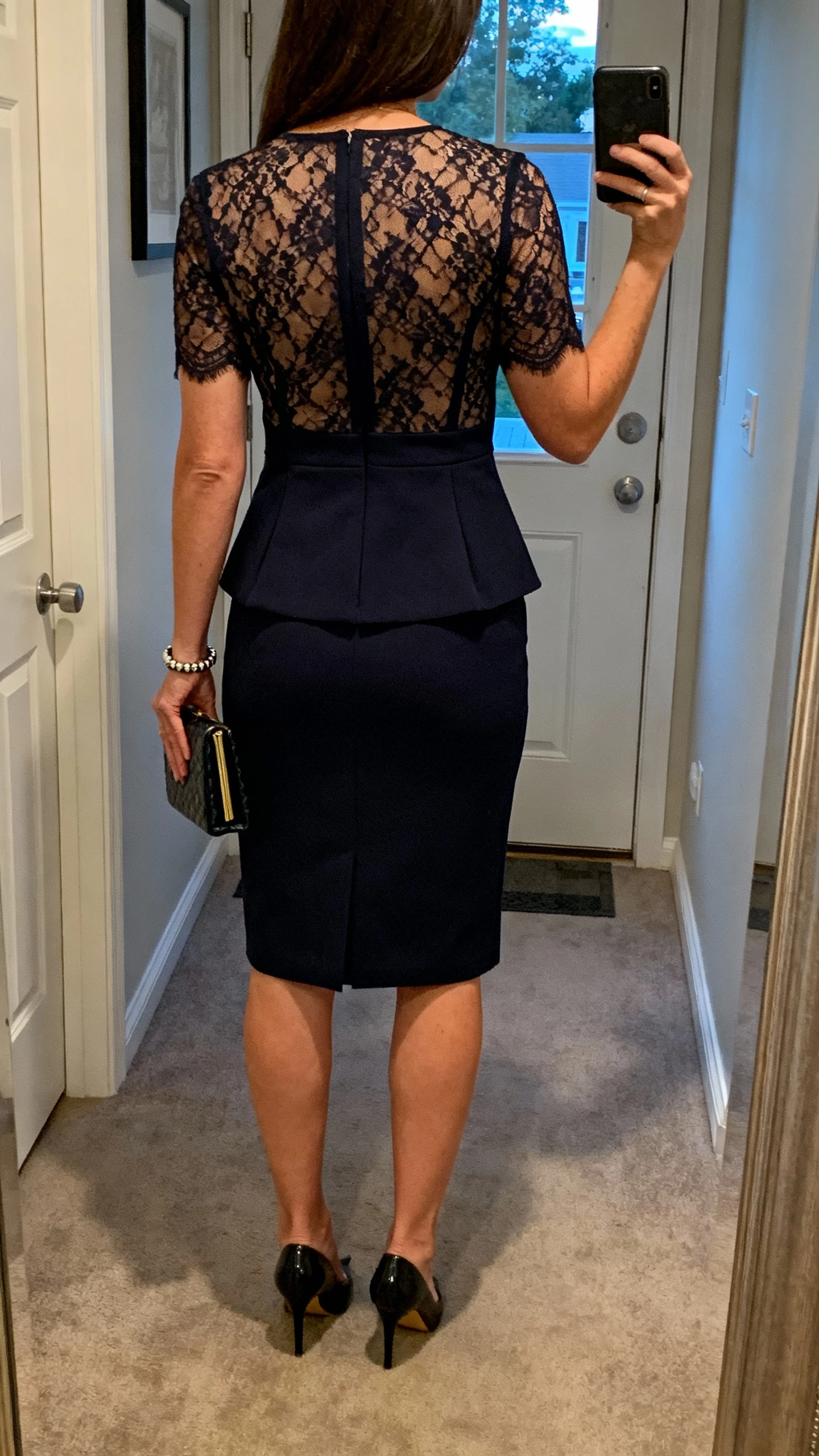 iPhone back-view selfie of a woman in a lace-and-tailoring midi sheath (lace overlay with clean tailored lines), classic pumps, understated bracelet, and a structured clutch, face not visible, hallway mirror by front door, early evening natural light, iPhone photo quality.