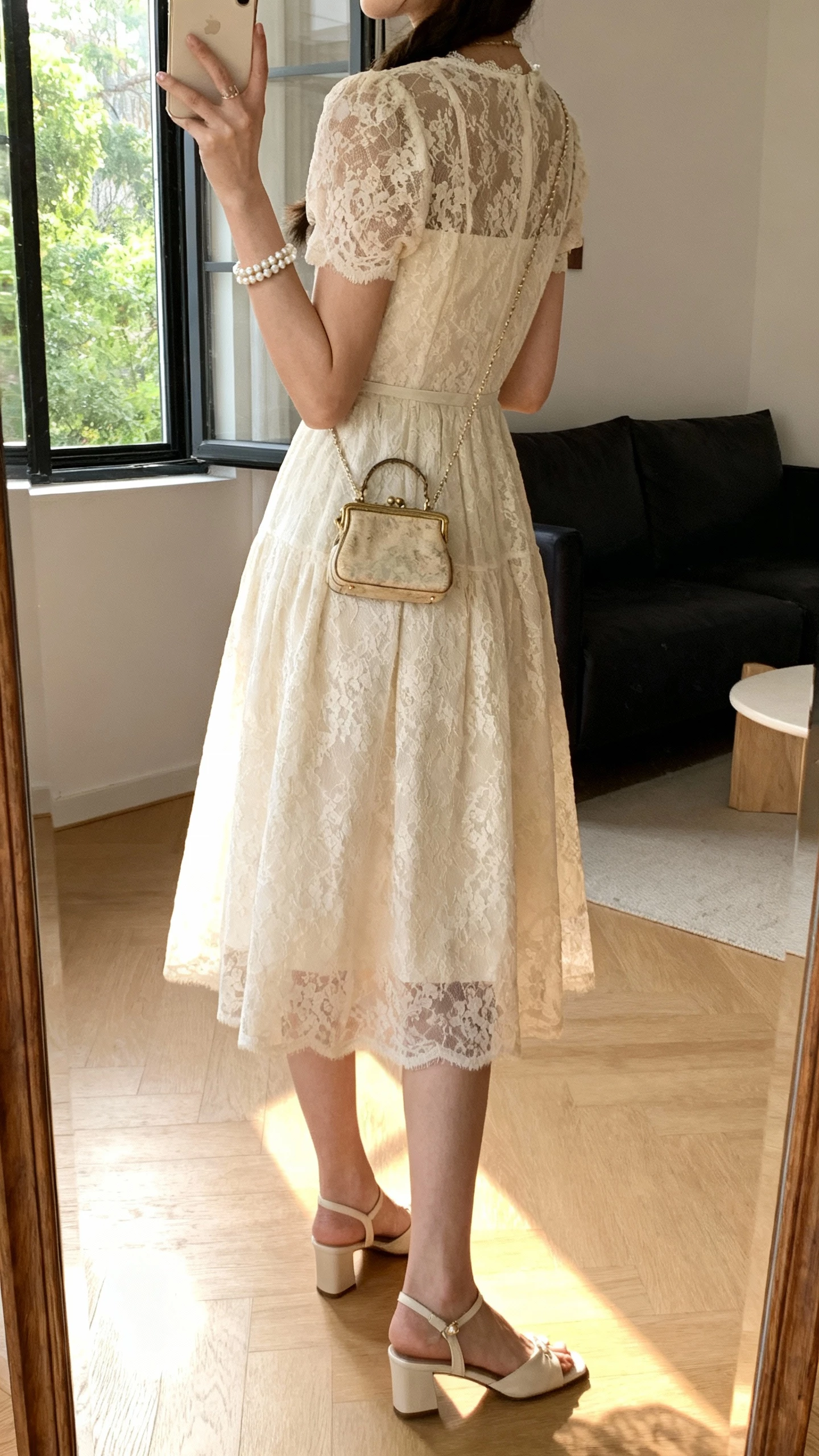 iPhone back-view selfie of a woman in a lace tea-length dress with slingback heels, pearl bracelet, vintage-style mini bag, face not visible, taken in a living room mirror with afternoon window light, casual iPhone shot