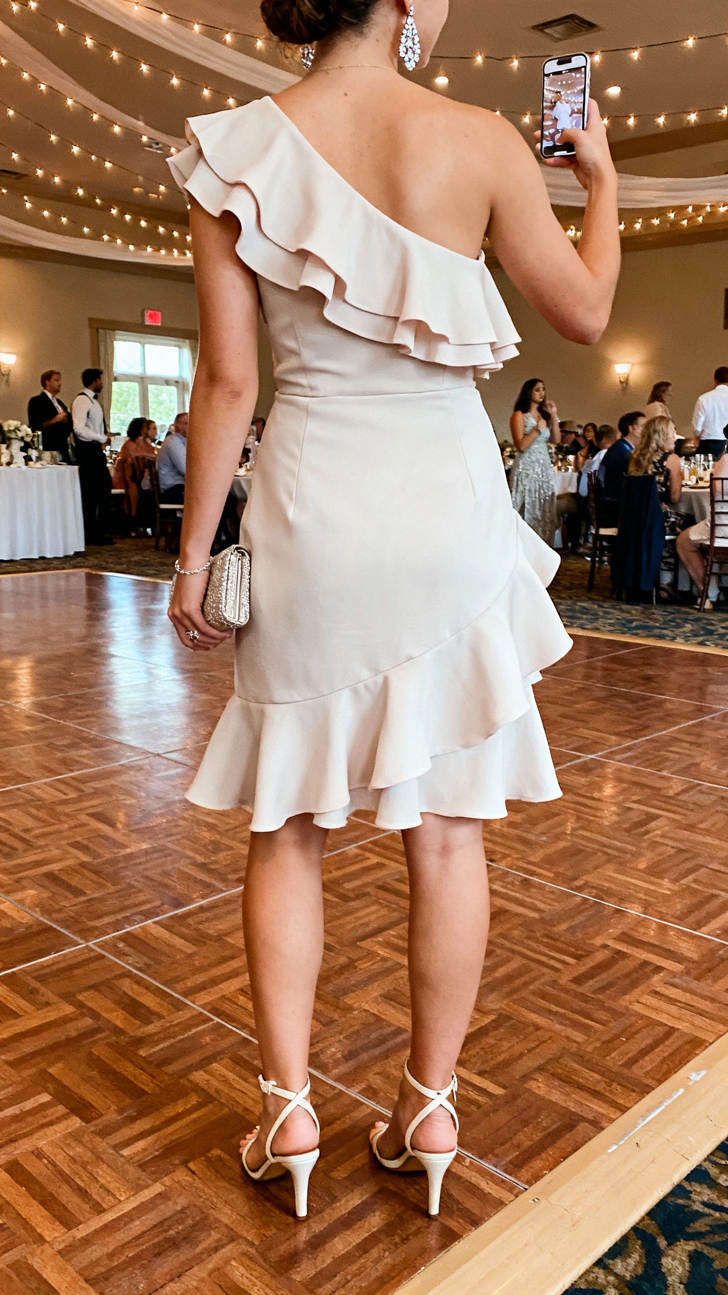 iPhone back-view selfie of a woman in a one-shoulder ruffle dress, ankle-strap heels, sparkly drop earrings, and a wristlet, face not visible, dance-floor edge in a reception hall, twinkle lights and natural iPhone quality