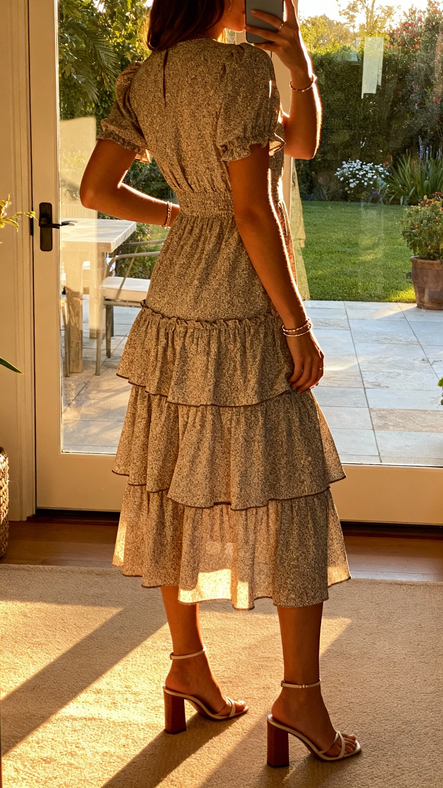 iPhone back-view selfie of a woman in a tiered ruffle midi dress with heeled sandals and simple bangles, face not visible, garden patio door reflection, late afternoon light, natural iPhone photo