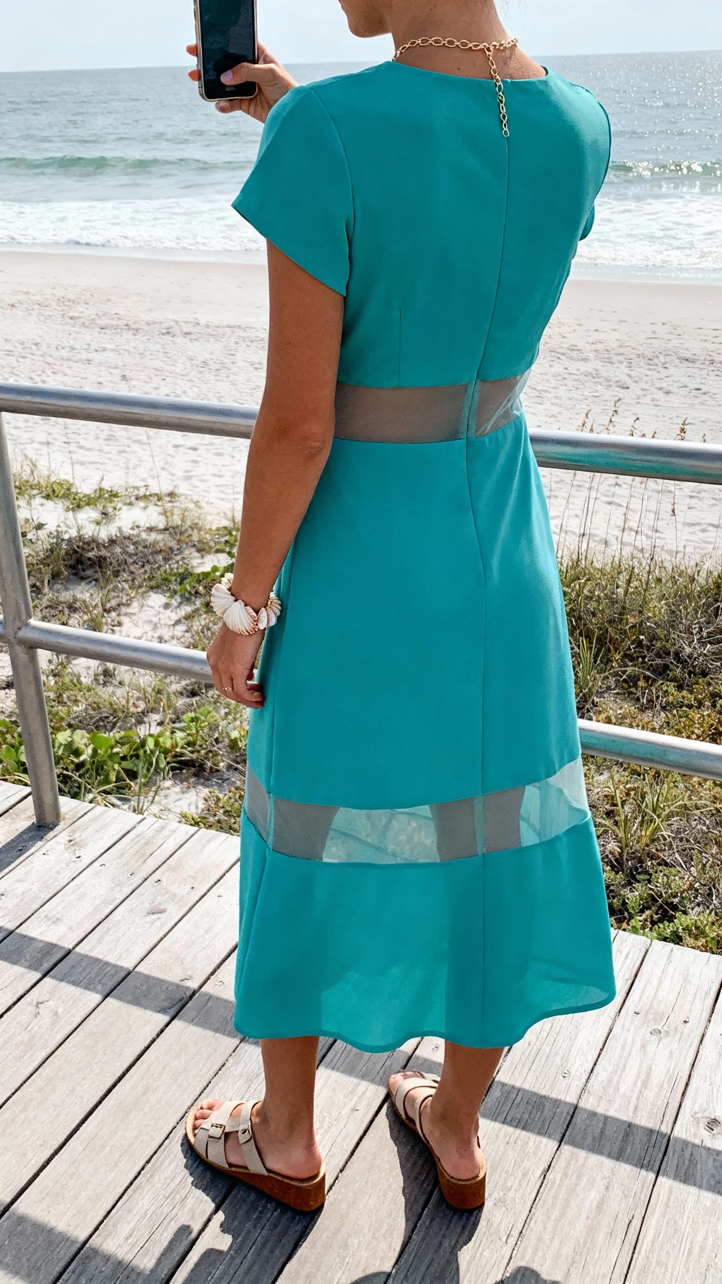 iPhone back-view selfie of a woman in an ocean-tone midi dress with tasteful sheer insets, dainty chain necklace, shell-inspired bracelet, low block sandals, face not visible, beachside boardwalk rail, soft coastal daylight, natural iPhone photo.