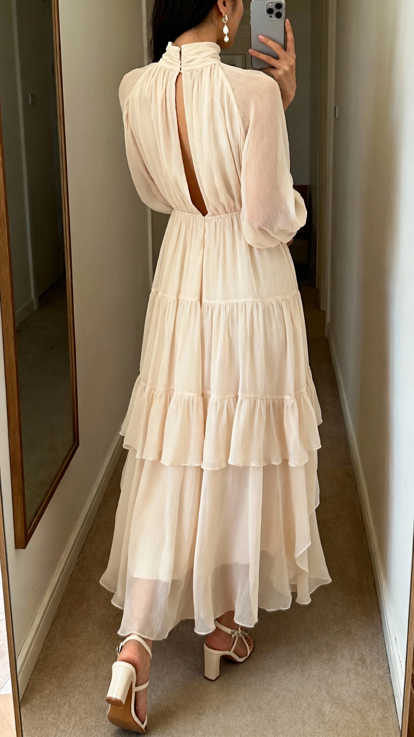 iPhone back-view selfie of a woman wearing a high-neck chiffon gown with a front keyhole, soft-flowing layers, heeled sandals, minimalist drop earrings, face not visible, corridor mirror with gentle indoor lighting, natural iPhone photo quality