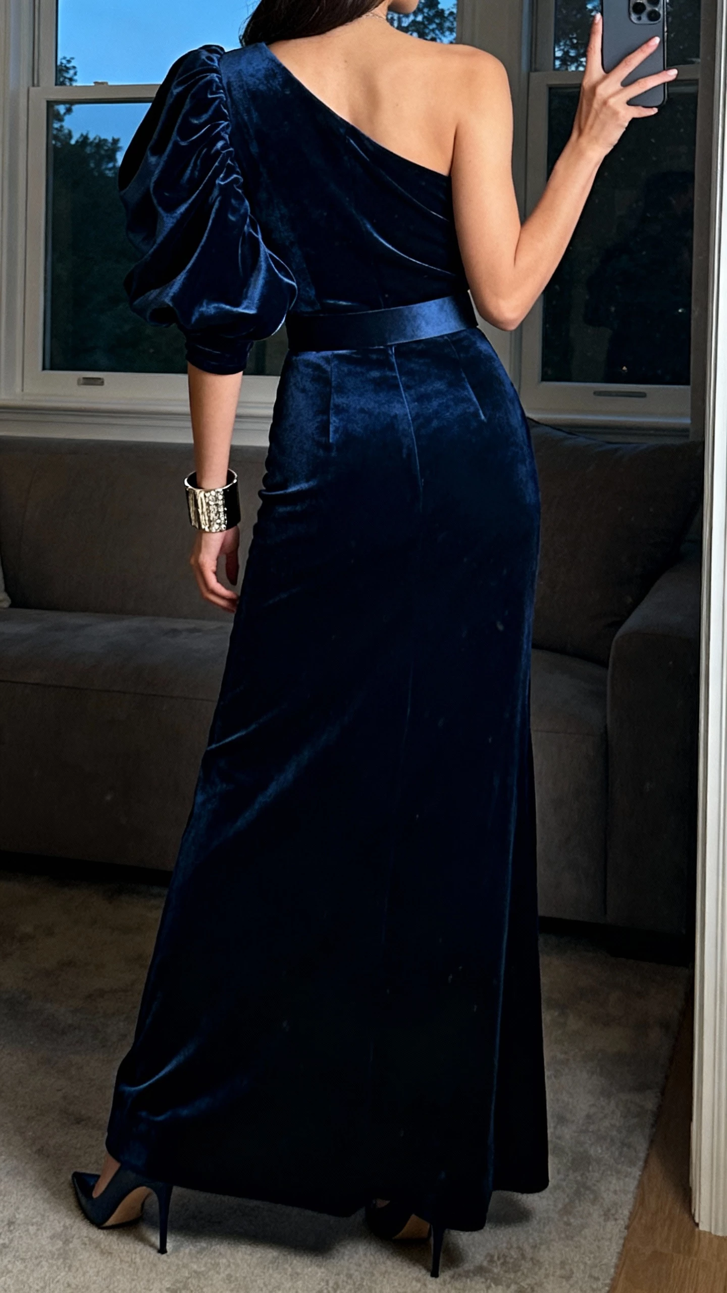 iPhone back-view selfie of a woman wearing a midnight blue one-shoulder velvet gown with dramatic puff sleeve and fitted waist, statement cuff bracelet, and pointed heels, face not visible, taken in a dim living room near a window, natural evening light, casual iPhone photo.