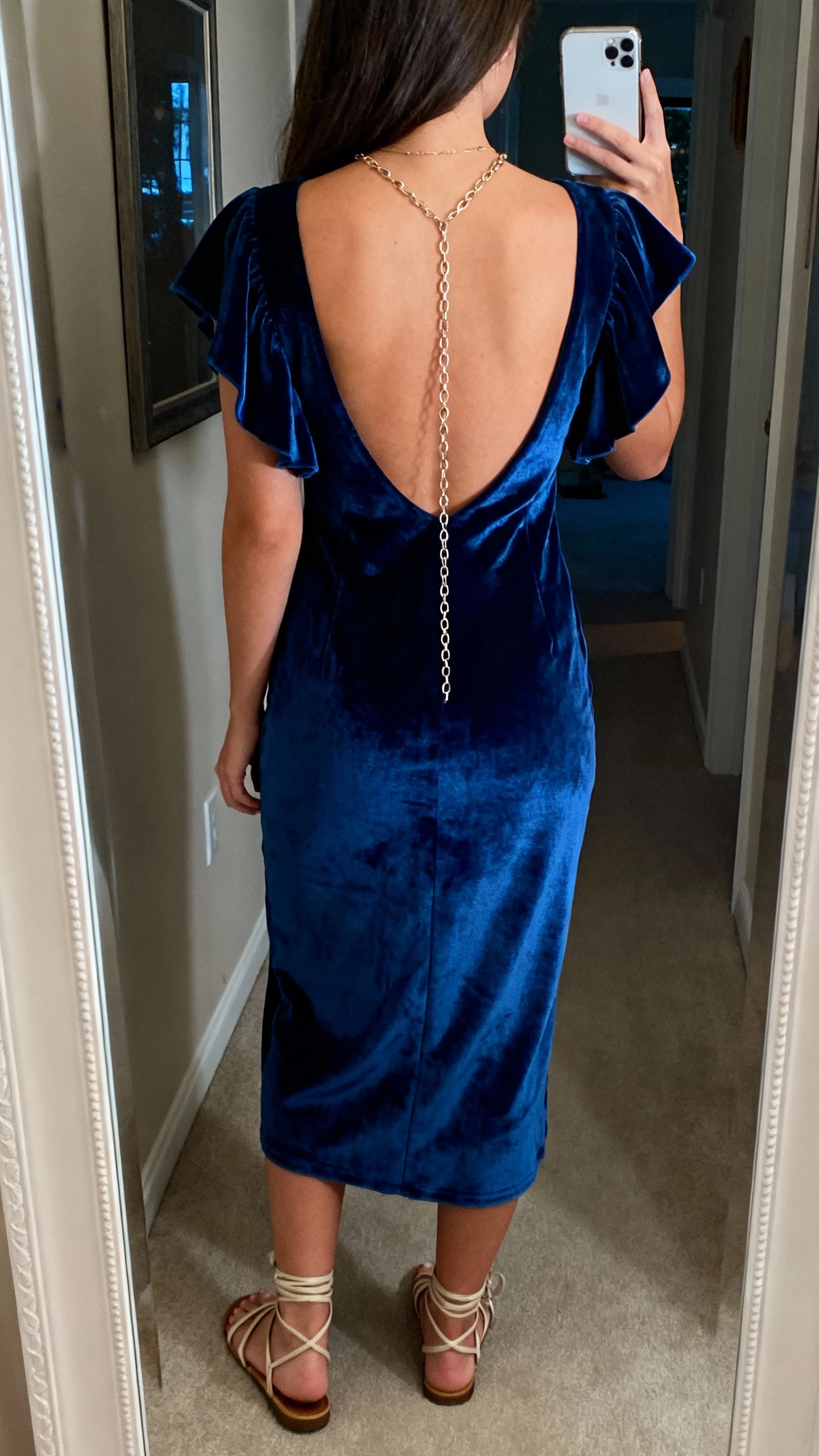 iPhone back-view selfie of a woman wearing a sapphire velvet dress with flutter sleeves and a low back, delicate chain necklace that drapes down, and strappy sandals, face not visible, hallway mirror before heading out, natural evening light, iPhone photo quality.