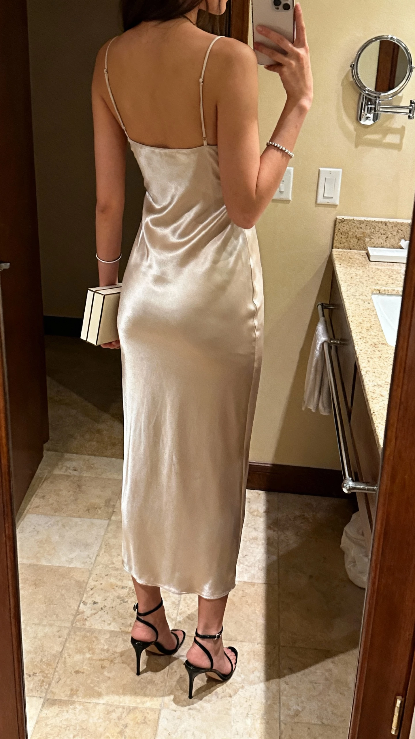 iPhone back-view selfie of a woman wearing a satin column dress with minimalist energy (straight silhouette, slim straps), sleek ankle-strap heels, thin bracelet, and a box clutch, face not visible, bathroom mirror, warm indoor lighting, natural iPhone photo quality.