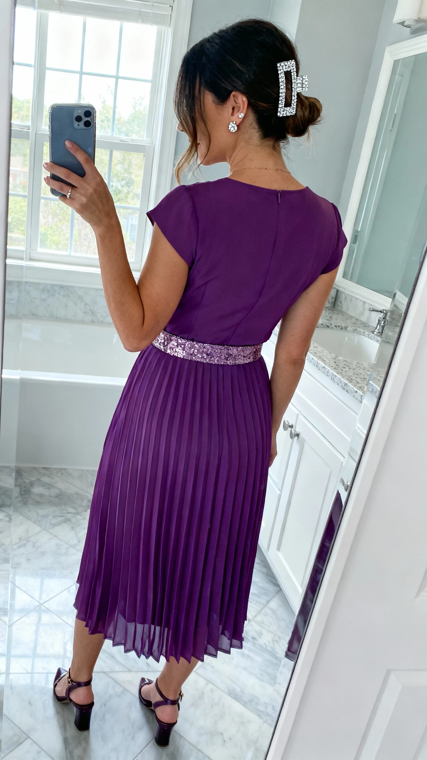 iPhone back-view selfie of a woman wearing an amethyst pleated midi dress with shimmer belt detail, pavé stud earrings, sparkly hair clip, and slingback heels, face not visible, taken in a bathroom mirror with cool morning light, natural iPhone quality.