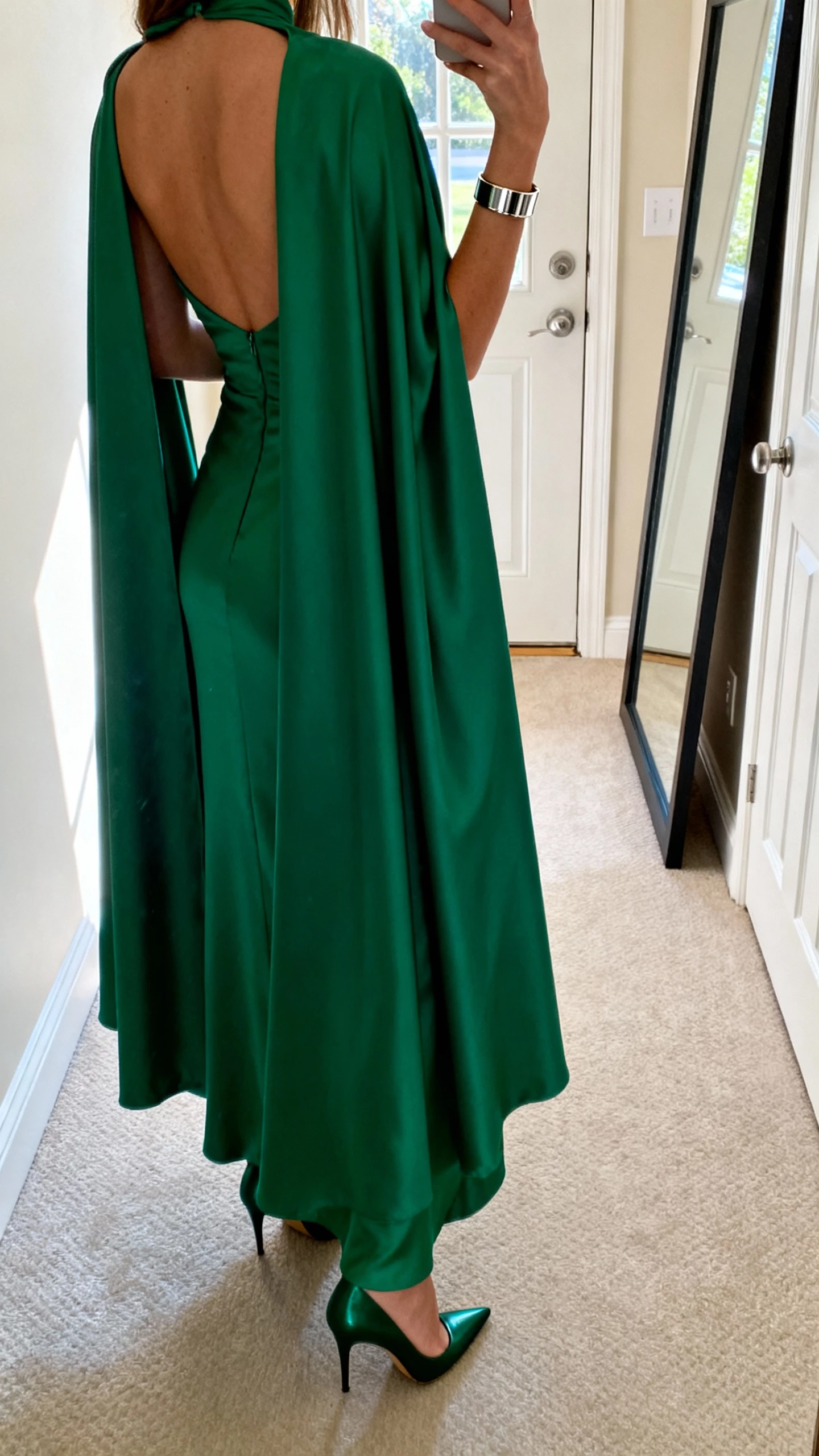 iPhone back-view selfie of a woman wearing an emerald cape-back gown for superhero glam, sleek bracelet, pointed heels, face not visible, hallway mirror near front door, daylight, natural iPhone photo.
