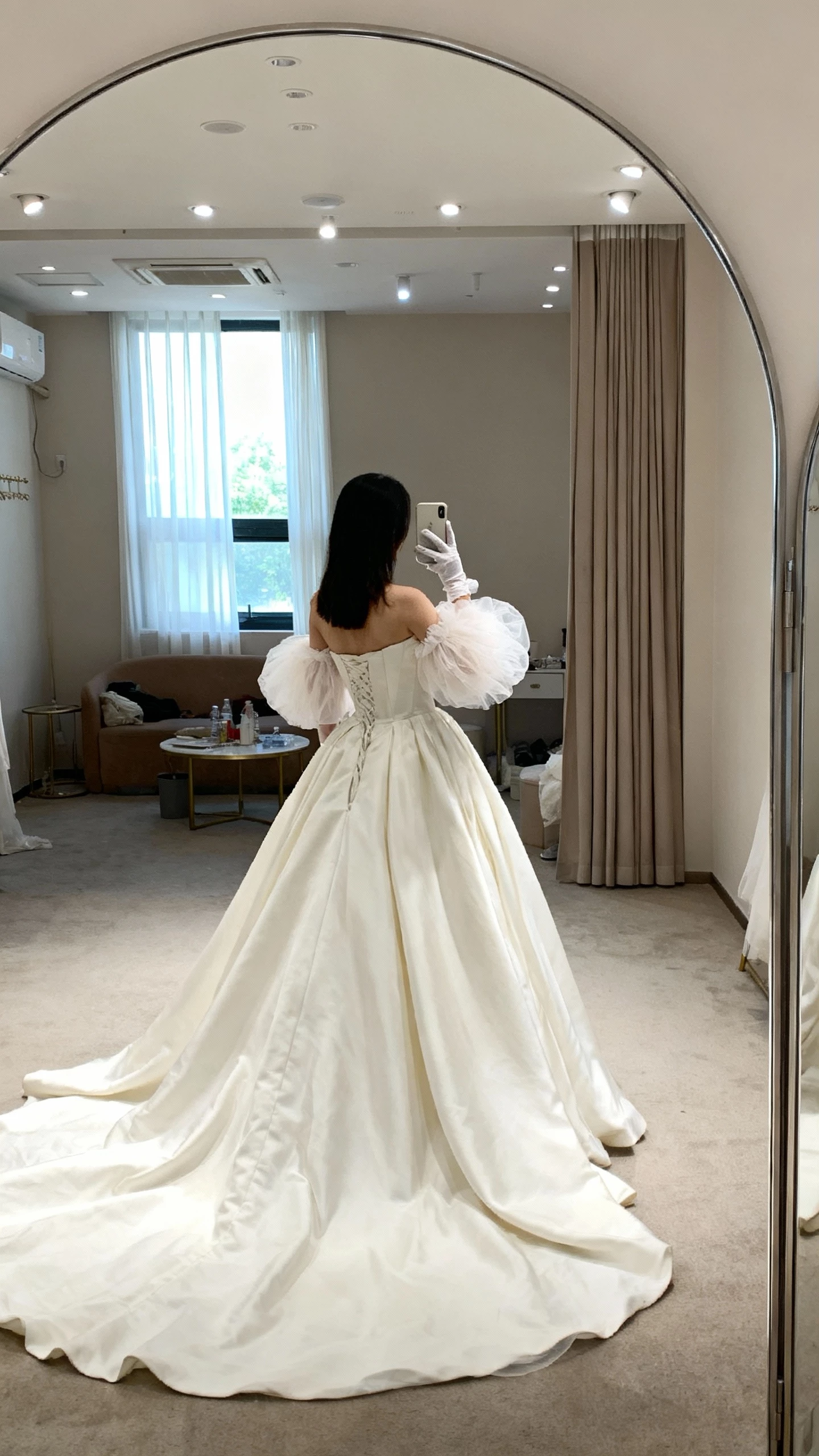 iPhone back-view selfie of a woman wearing detachable puff-tulle gloves with a classic princess gown, long train visible, face not visible, venue bridal suite mirror, gentle window light, natural iPhone photo quality