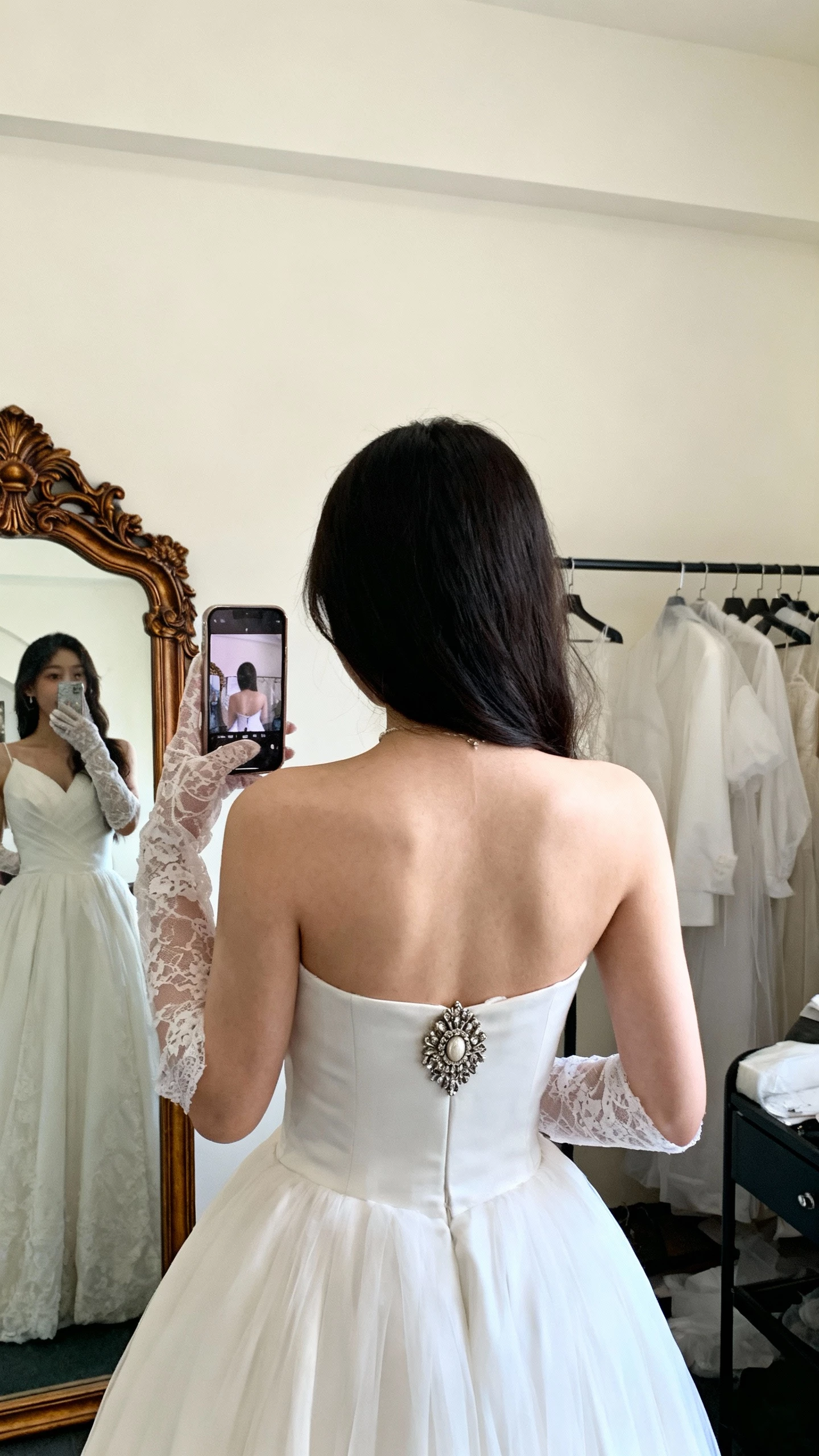 iPhone back-view selfie of a woman wearing lace gloves, an heirloom brooch pinned at the portrait neckline of her wedding dress, face not visible, dressing room with vintage mirror, soft indoor lighting, natural iPhone photo quality