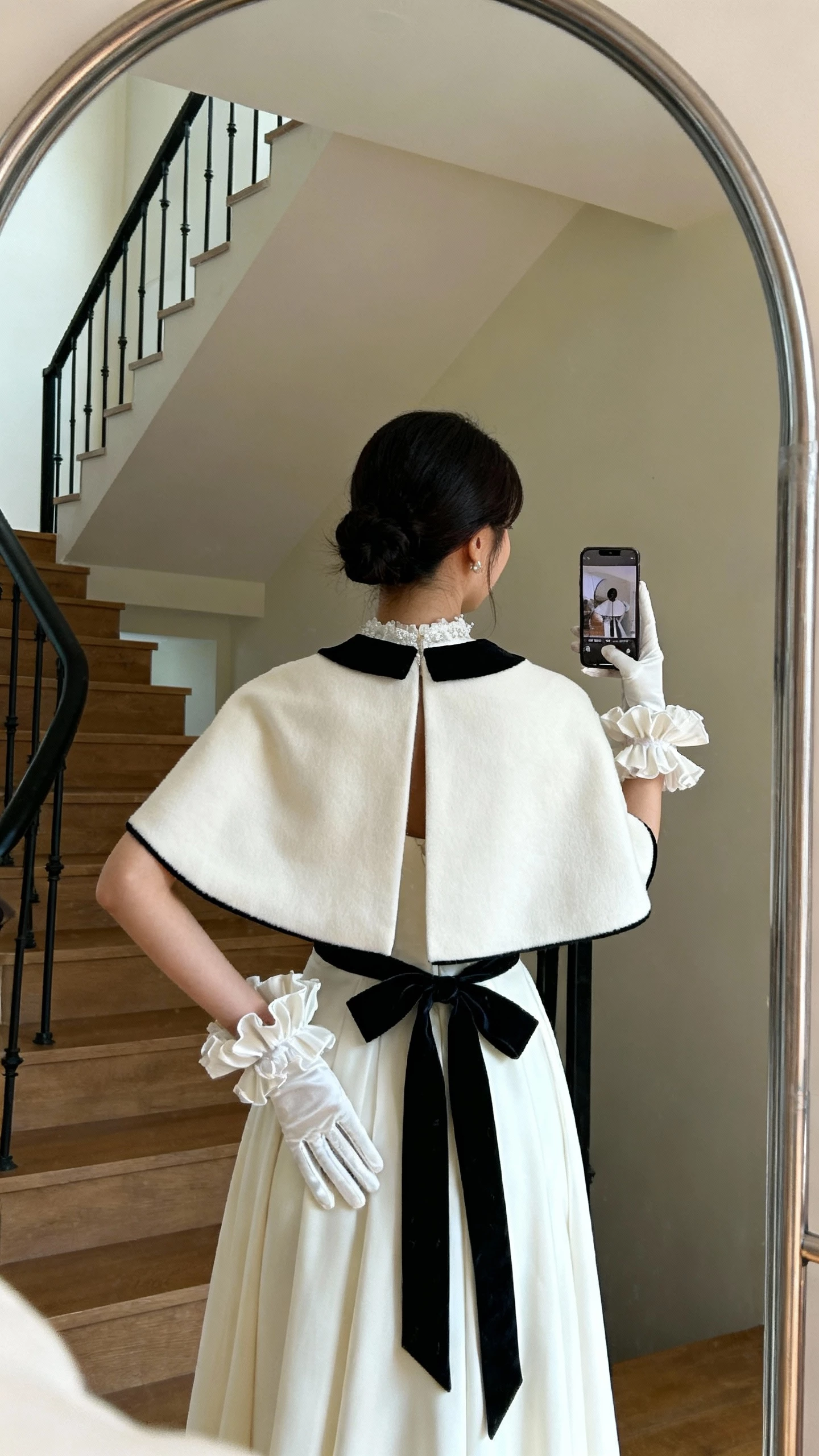 iPhone back-view selfie of a woman wearing ruffled cuff gloves, a statement cape, and velvet ribbon details on her bridal look, face not visible, staircase mirror, indoor ambient lighting, casual iPhone photo