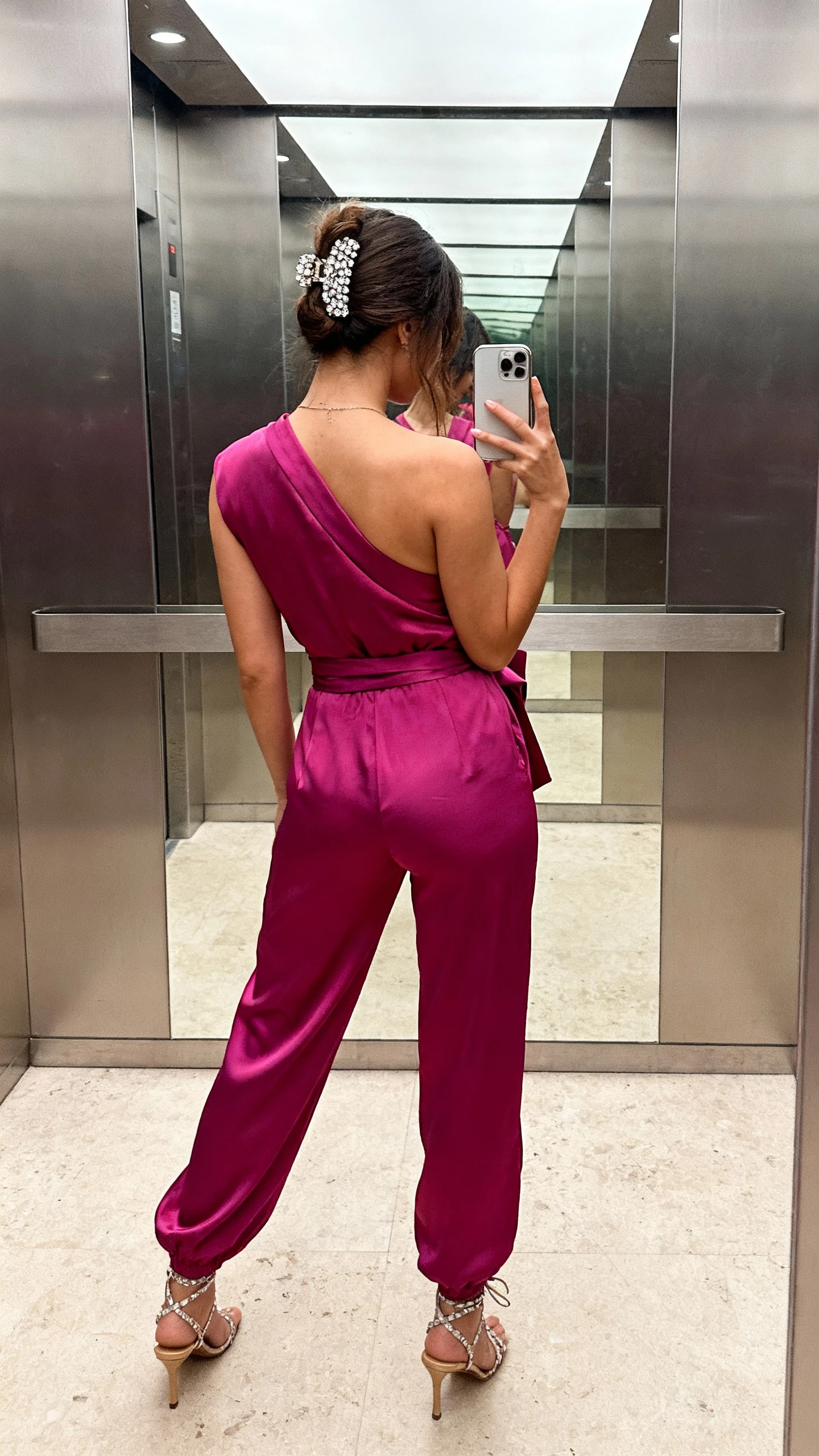 iPhone back-view selfie showing a woman in a jewel-tone wrap jumpsuit with a bejeweled hair clip, strappy heels, face not visible, elevator mirror, soft overhead lighting, casual iPhone shot.