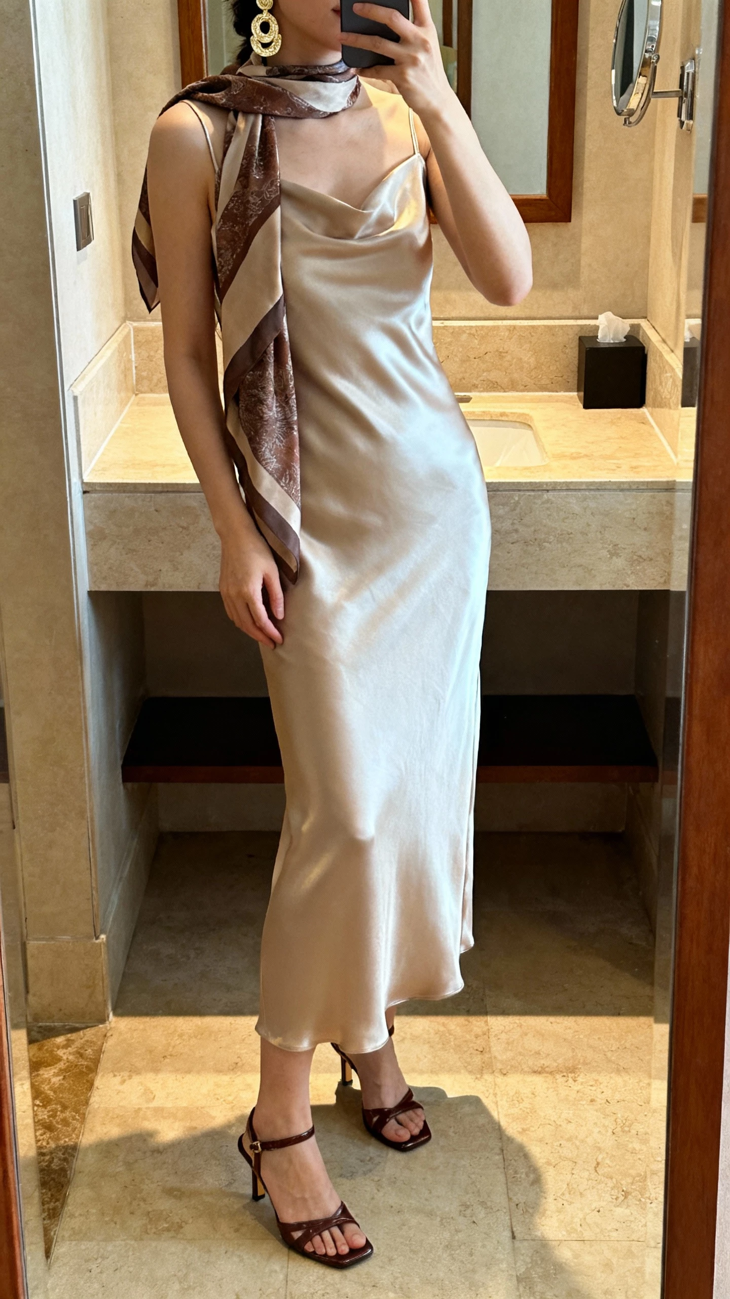 iPhone bathroom mirror selfie of a woman in a satin column dress with a coordinating scarf and statement earrings, sleek heeled sandals, face not visible, warm indoor lighting, natural iPhone photo.