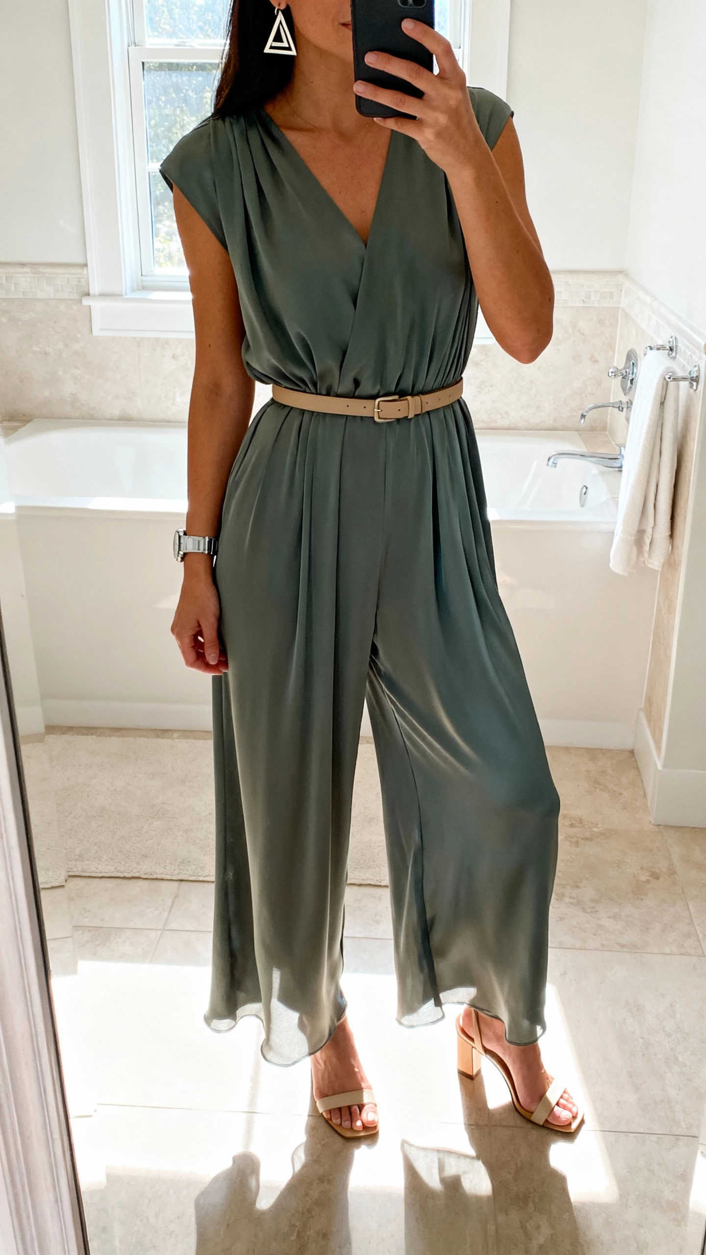 iPhone bathroom mirror selfie of a woman in a wide-leg jumpsuit with liquid drape, slim belt, heeled sandals, geometric earrings, wristwatch, face not visible, bright overhead and window light, natural iPhone photo quality