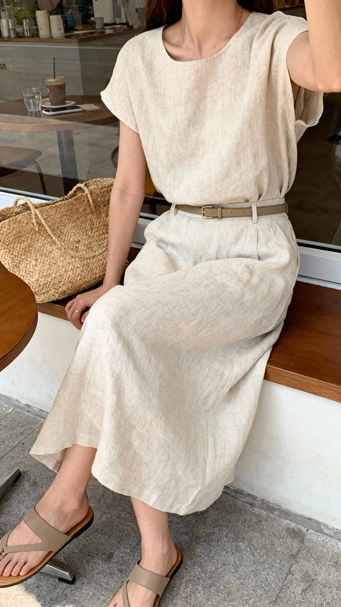 iPhone cafe table selfie cropped at neck showing a woman in a linen-blend midi set (matching top and skirt), slide sandals, a slim belt, and a straw tote, face not visible, seated by a window in a cafe, soft morning light, natural iPhone photo quality.