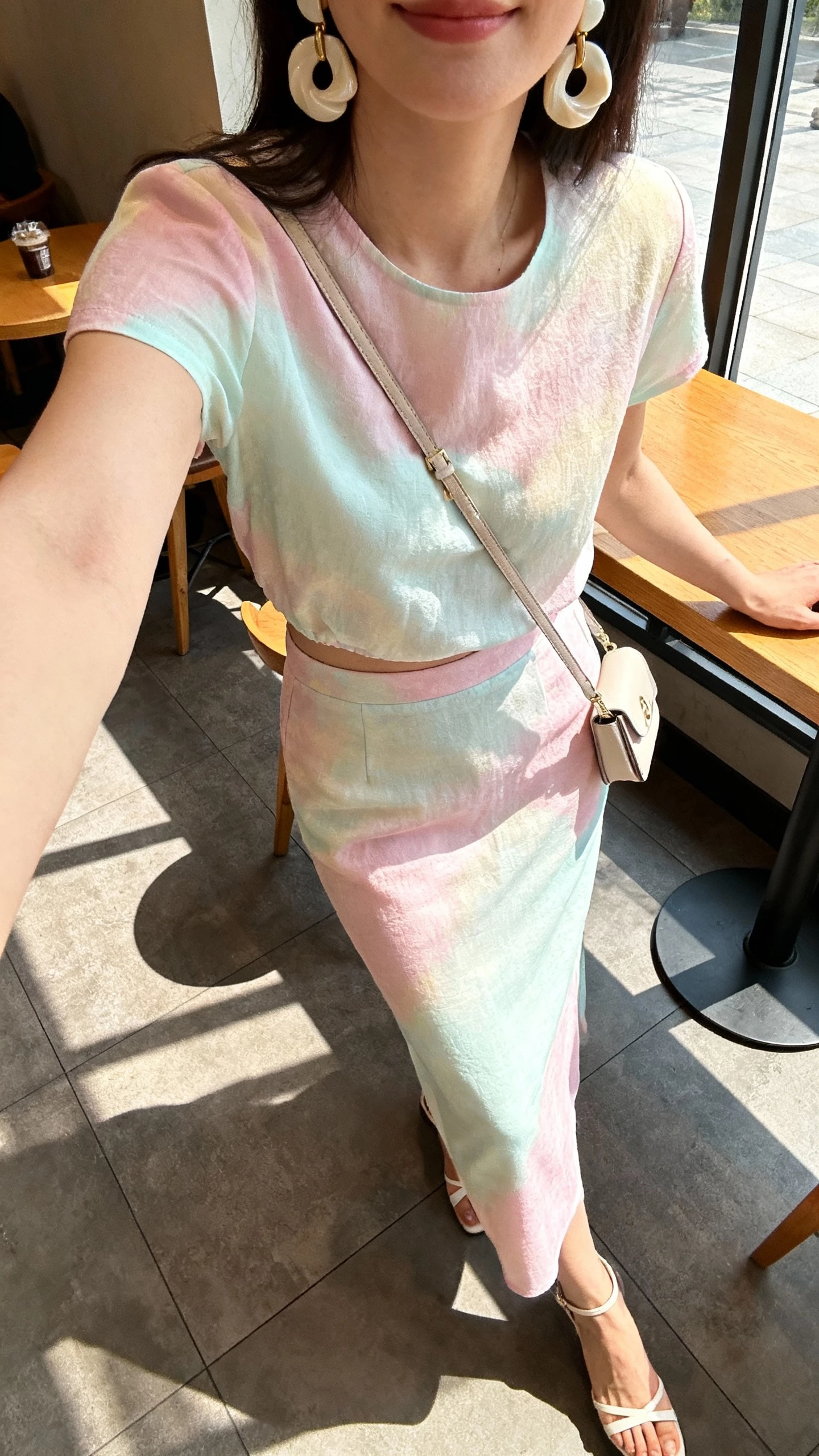 iPhone cafe table selfie cropped at neck showing a woman wearing a pastel co-ord skirt set with sculptural earrings, slim ankle-strap heels, small crossbody, face not visible, sunny cafe window light, casual iPhone quality