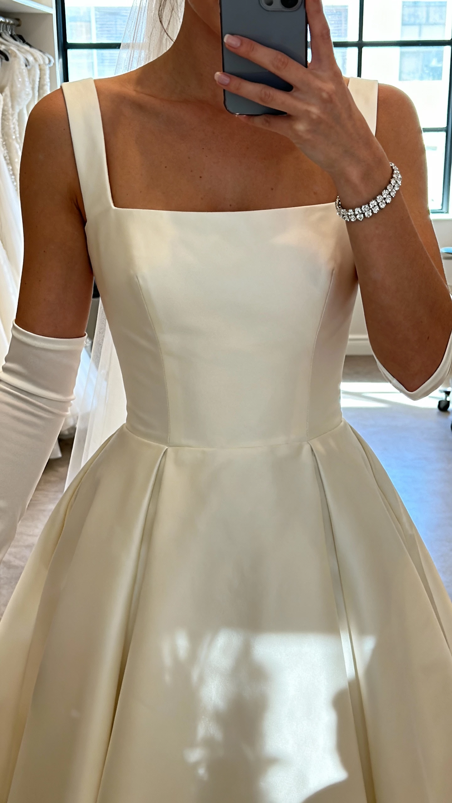iPhone chest-up selfie of a woman wearing elbow-length gloves, a diamond tennis bracelet, and a square neckline wedding dress, face not visible, bridal suite window light, natural iPhone photo quality
