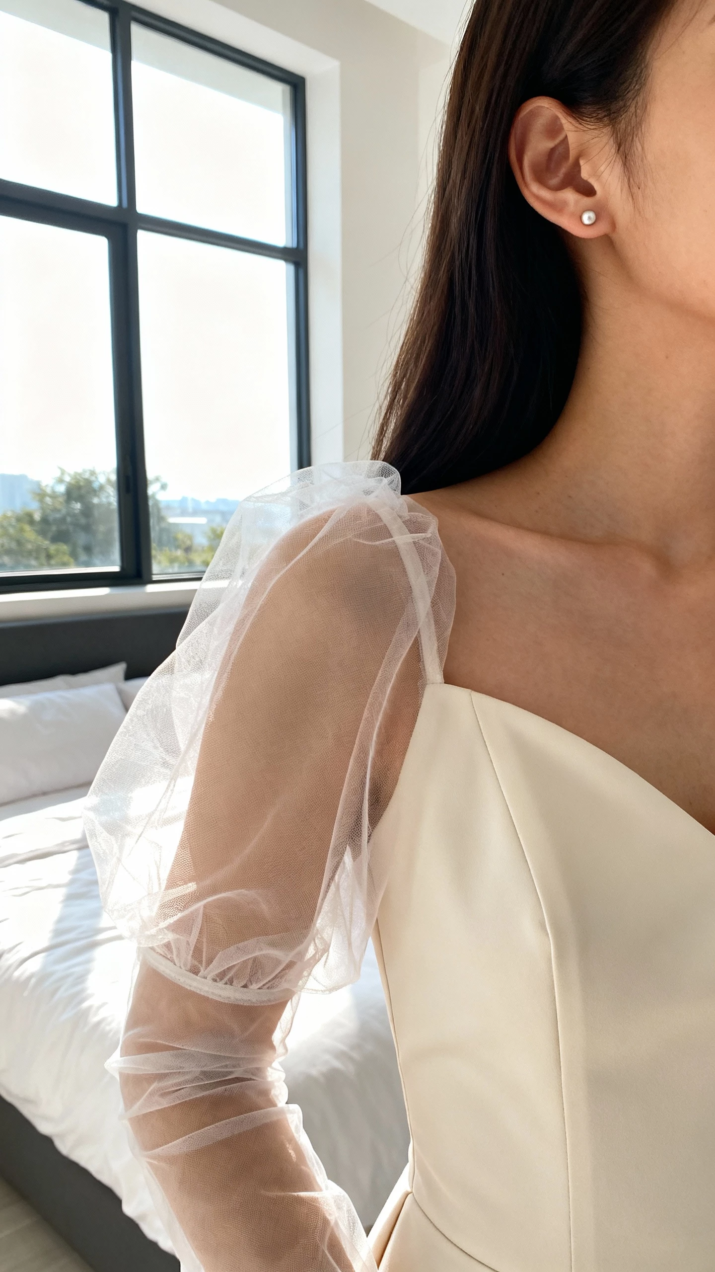 iPhone close crop at shoulders showing a woman wearing sheer tulle gloves with a minimalist crepe gown, simple stud earrings, face not visible, standing by a bright window in a modern bedroom, morning light, natural iPhone photo quality