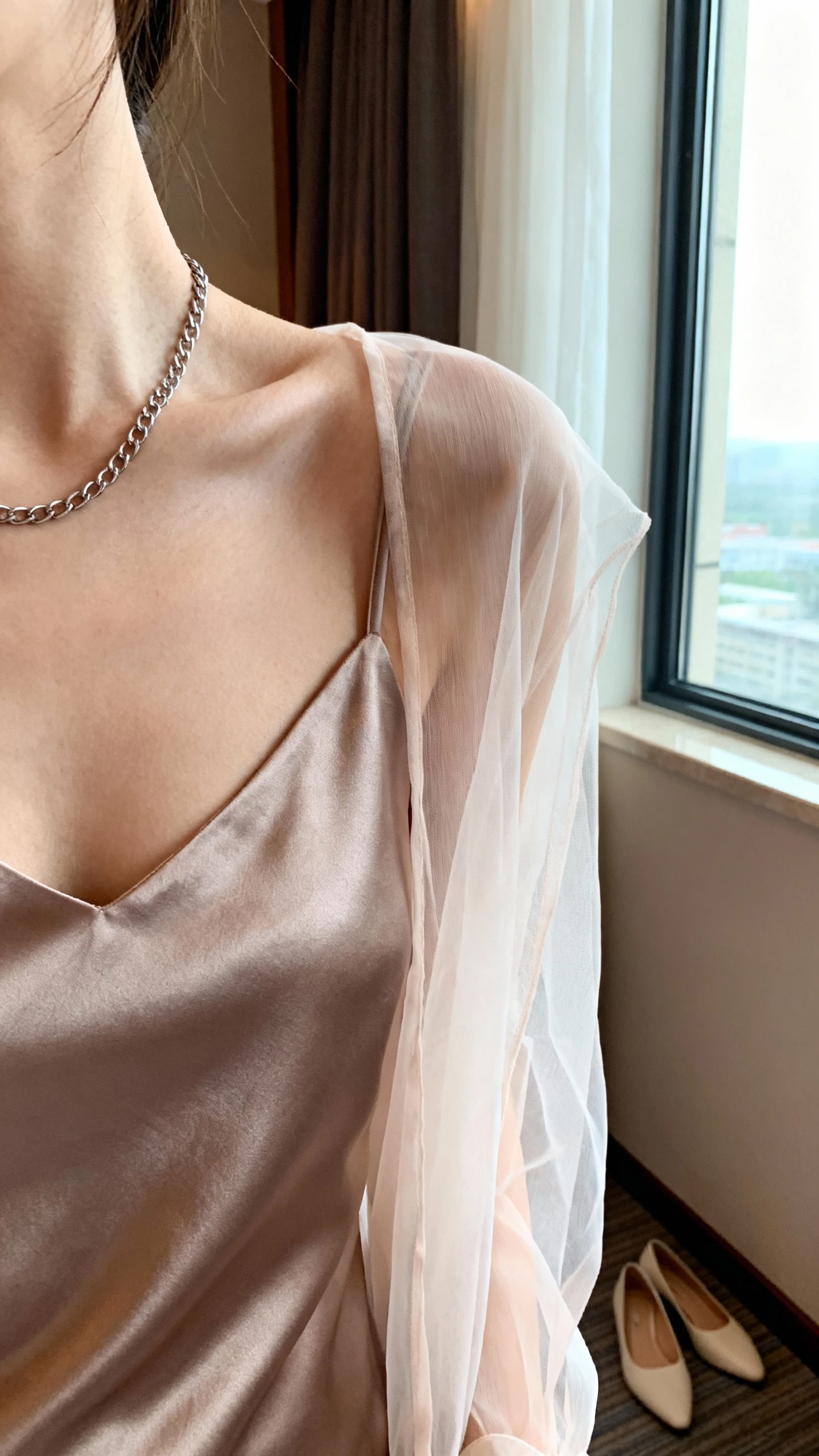 iPhone close crop at shoulders showing a woman wearing a silk slip dress layered with a sheer organza topper, delicate chain necklace, minimalist heels, face not visible, taken near a window in a hotel room, soft natural light, iPhone photo quality