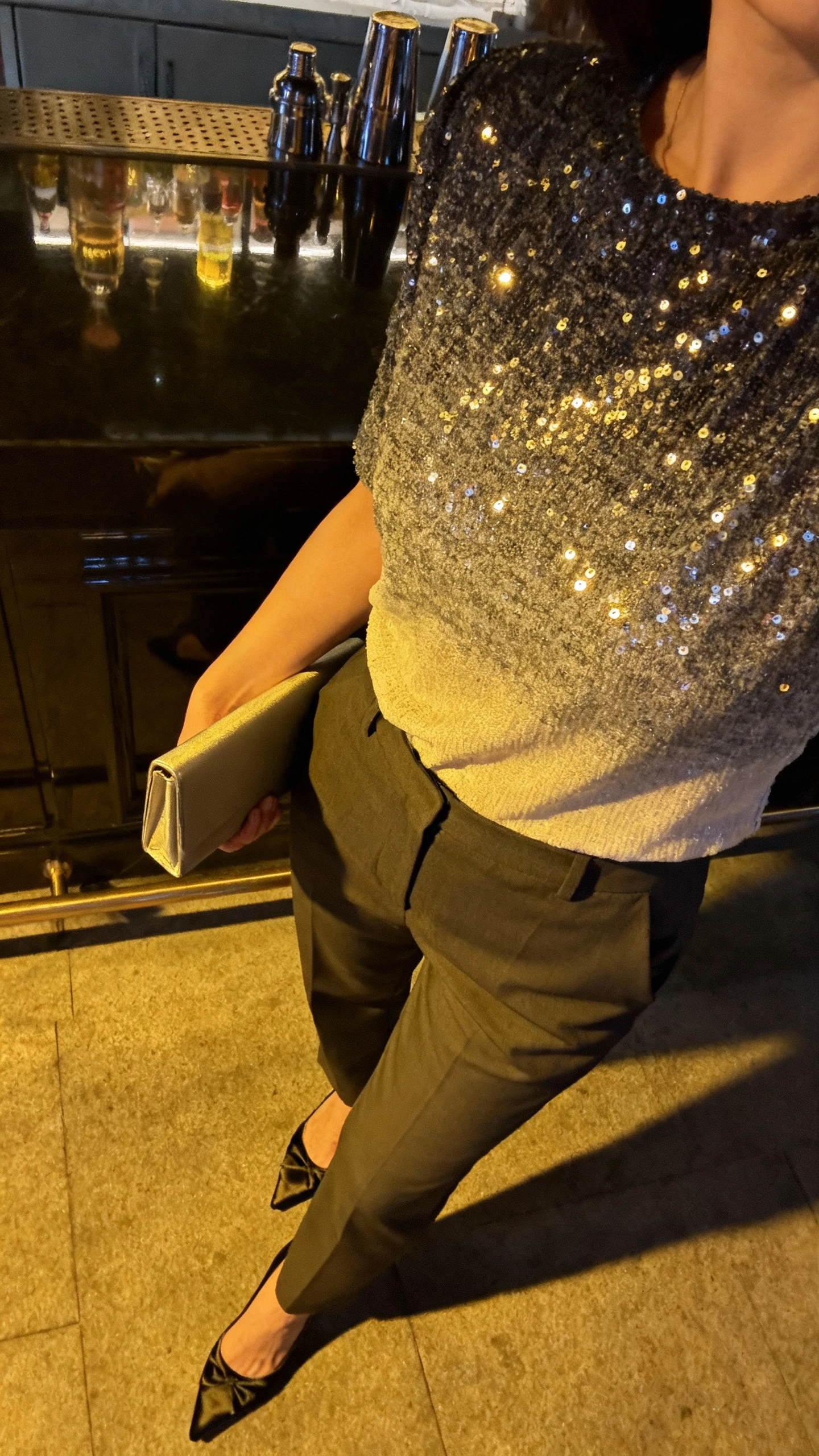 iPhone cropped-at-shoulders selfie of a woman in an ombre sequin top, tailored pants, and satin heels, simple clutch, face not visible, chic bar table reflection, evening ambient light, natural iPhone aesthetic.