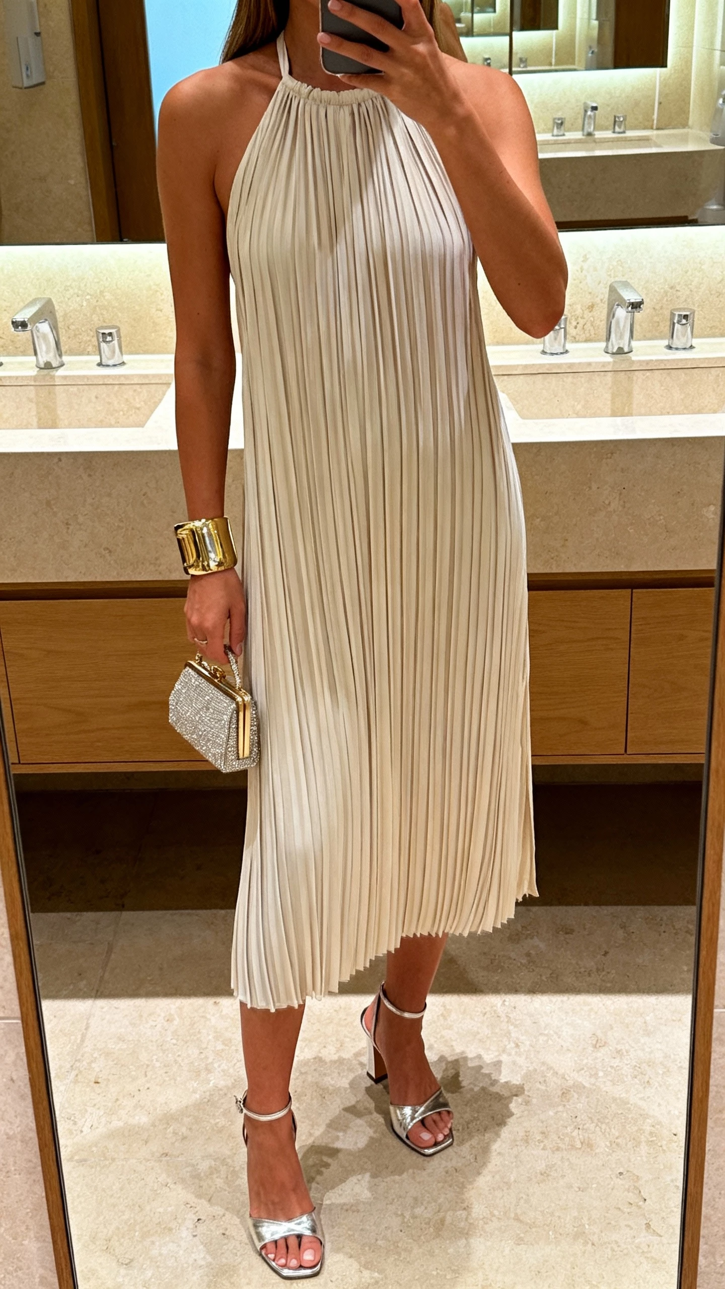 iPhone cropped-at-shoulders selfie of a woman wearing a pleated halter dress, metallic heeled sandals, statement cuff bracelet, and a petite evening bag, face not visible, chic restroom mirror at a venue, indoor ambient lighting, natural iPhone photo