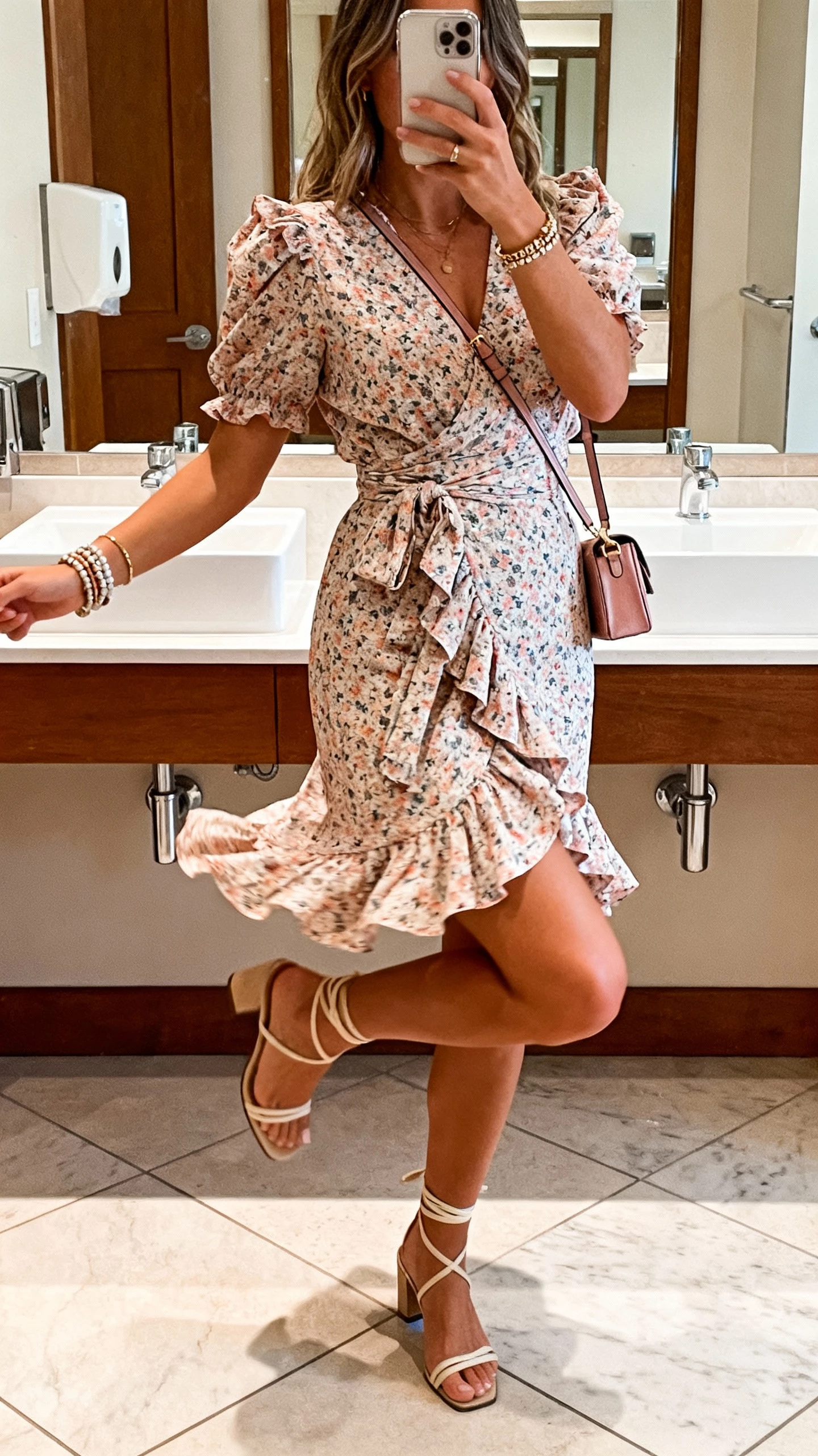 iPhone dance-floor bathroom mirror selfie showing a woman in a ruffled wrap floral dress with movement, ankle-strap heels, layered bracelets, and a mini shoulder bag, face not visible, warm indoor lighting, casual iPhone aesthetic.