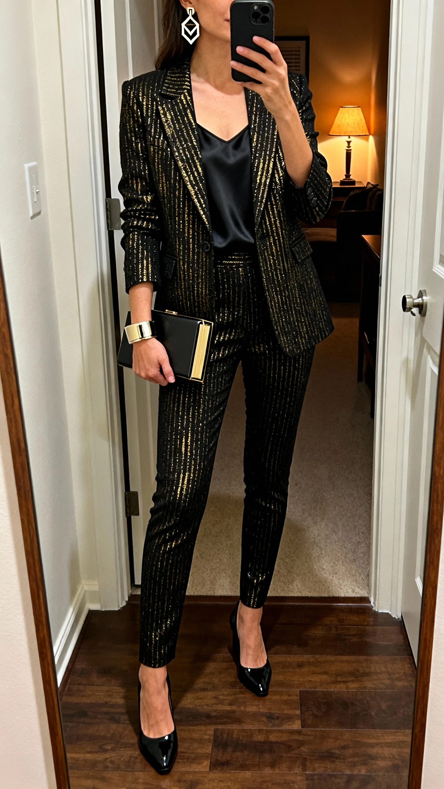iPhone full-length mirror selfie of a woman in a single-breasted blazer with black-gold metallic thread, slim matching trousers, minimalist black silk tank, black patent pumps, geometric statement earrings with a cuff bracelet, box clutch; apartment entryway, face not visible (cropped at neck), evening lamp light, natural iPhone photo quality.