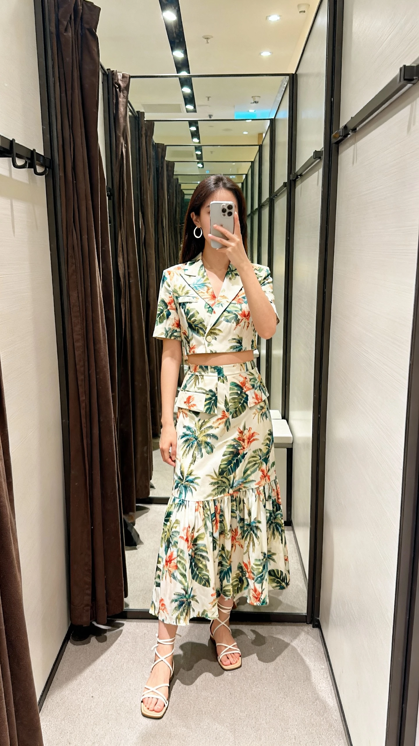 iPhone full-length mirror selfie of a woman wearing a tropical tailored set: cropped top and flowy skirt, strappy sandals, simple hoop earrings, face not visible, boutique dressing room mirror, overhead indoor lighting, casual iPhone photo.