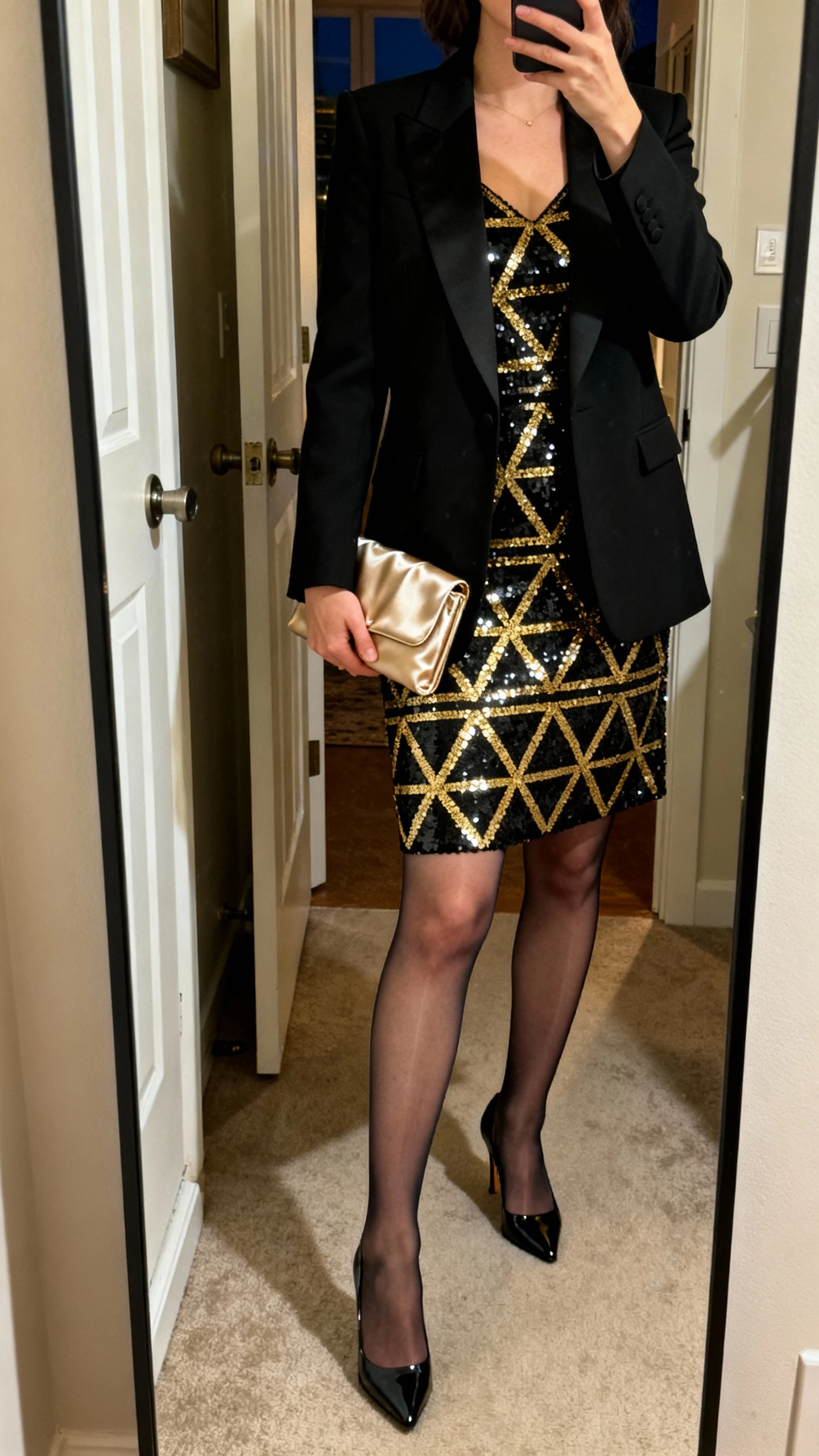 iPhone full-length mirror selfie of a woman wearing a black-and-gold geometric sequin dress paired with a tailored black tuxedo blazer, satin clutch, sheer tights, and patent heels, face not visible, apartment entry mirror, natural evening light, iPhone photo quality.