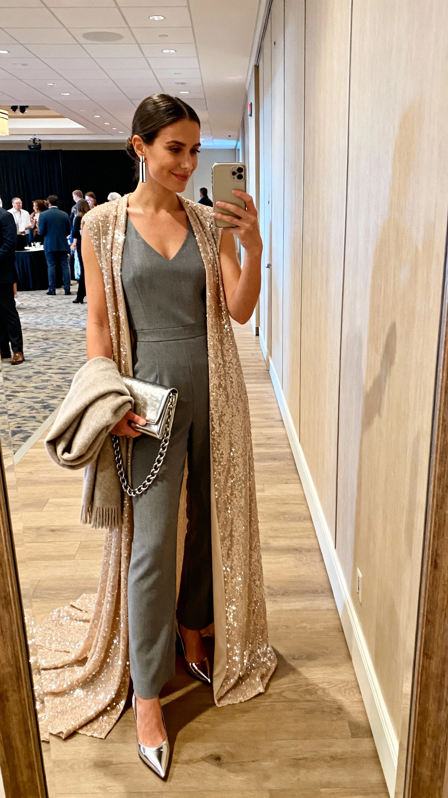 iPhone hallway mirror selfie of a woman in a graphite sleeveless crepe jumpsuit under a floor-sweeping champagne sequined duster, pointed metallic pumps, sleek bar earrings, metallic chain-strap clutch, cashmere wrap folded in hand; face not visible, event venue corridor, warm indoor light, natural iPhone photo.