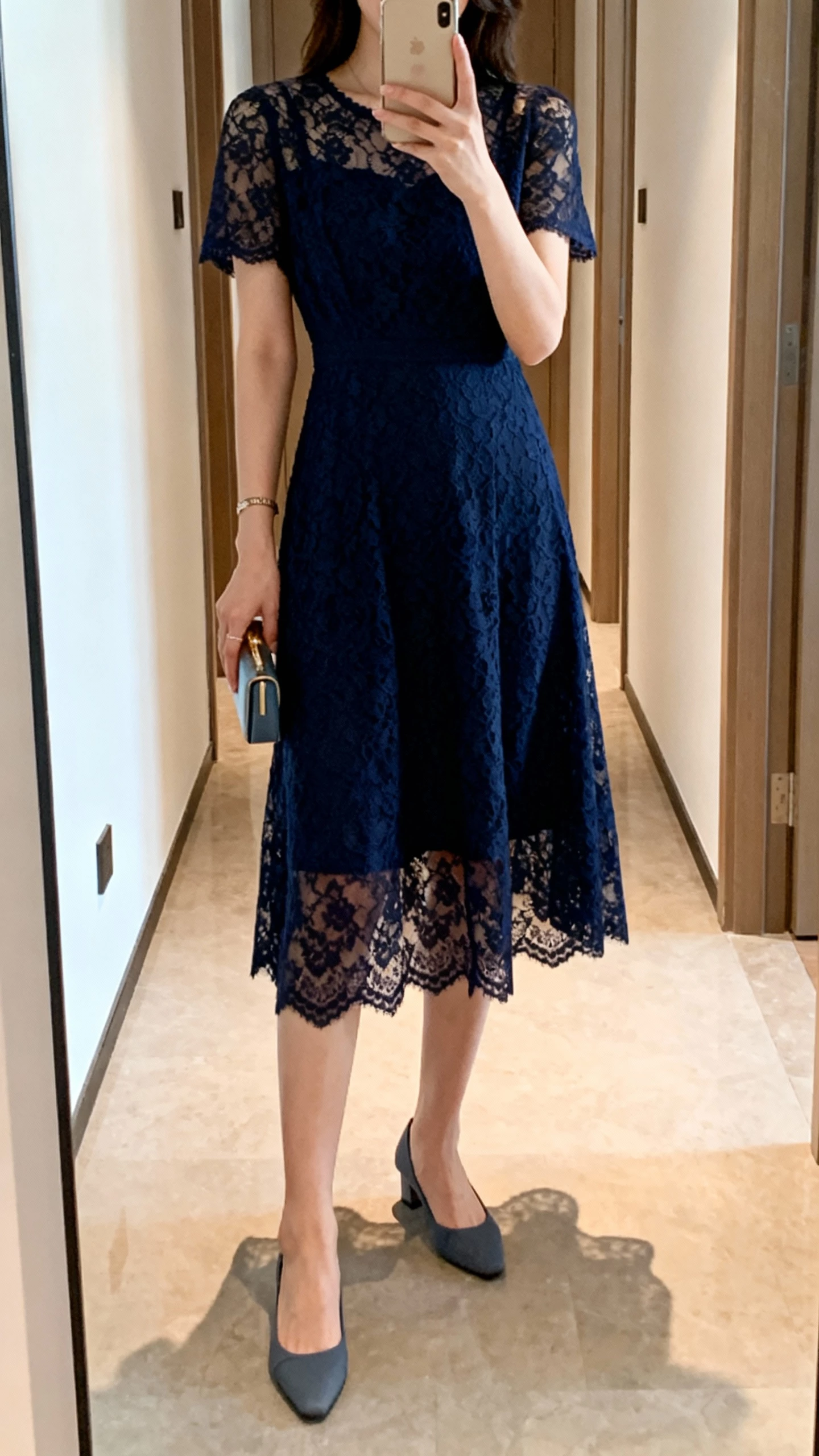 iPhone hallway mirror selfie of a woman in a navy lace midi dress with minimalist heels, tiny clutch, face not visible, cropped at shoulders, warm indoor lighting, casual iPhone photo