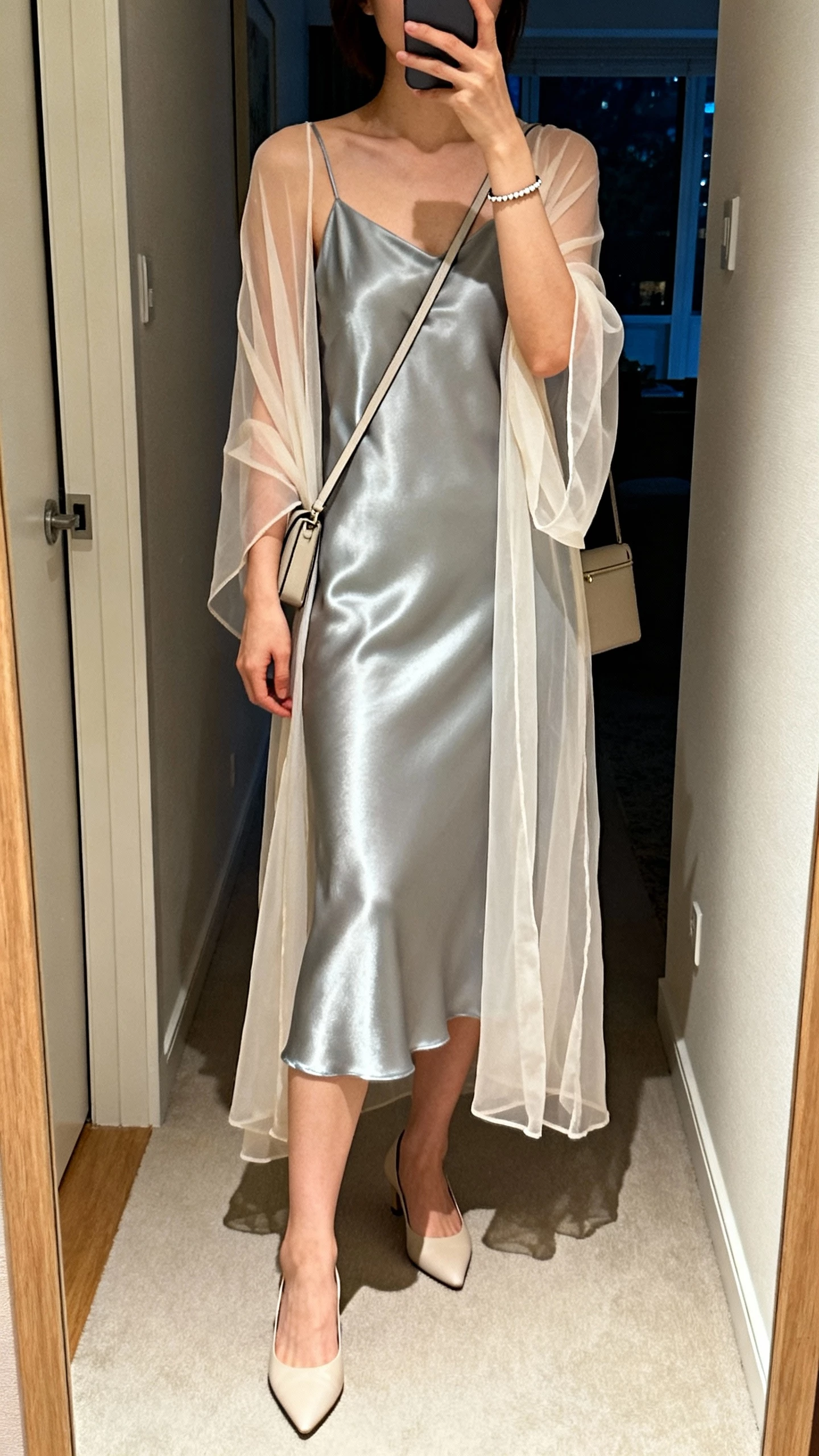 iPhone hallway mirror selfie of a woman in a satin slip dress layered with a weightless sheer duster, minimalist heels, slim shoulder bag, simple bracelet, face not visible, soft indoor evening lighting, natural iPhone photo quality