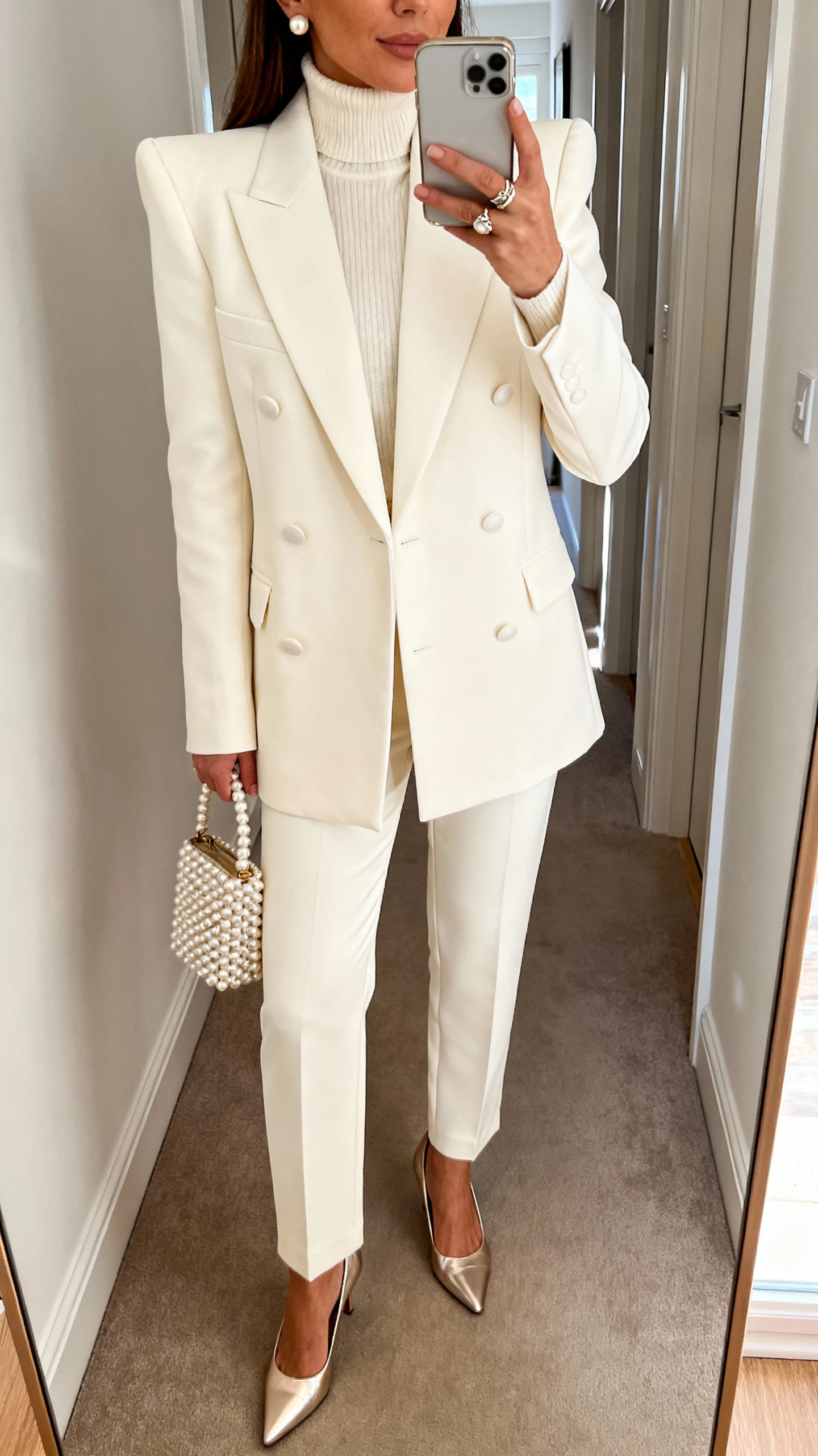 iPhone hallway mirror selfie of a woman in a winter-white double-breasted cream blazer with strong shoulders, matching high-waisted straight-leg trousers, ivory cashmere turtleneck, champagne metallic heels, pearl studs with a statement ring, mini pearlized top-handle bag; face not visible (cropped at shoulders), soft morning light, natural iPhone photo quality.