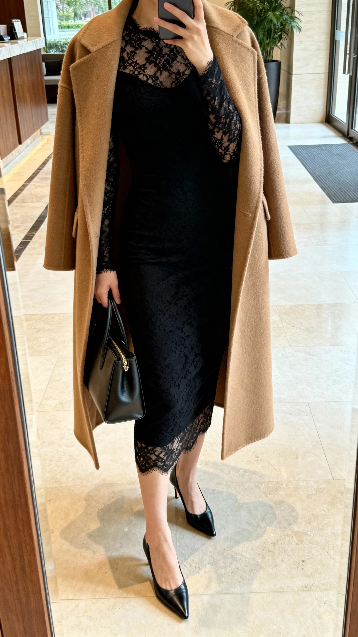iPhone hallway mirror selfie of a woman wearing a long-sleeve black lace sheath dress under a dramatic camel wool coat, black pointed pumps, and structured tote; face not visible, cropped at neck, lobby mirror, morning natural light, iPhone photo quality.