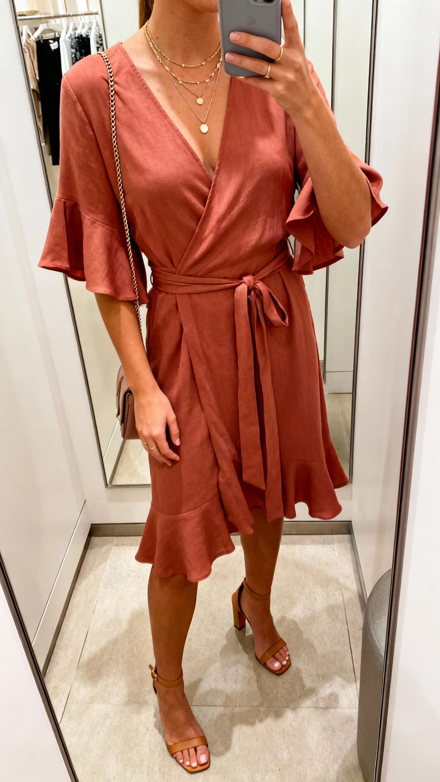 iPhone mirror selfie cropped at neck of a woman wearing a romantic wrap dress (tie waist, flutter sleeves), heeled sandals, layered delicate necklaces, and a small crossbody, face not visible, boutique dressing-room mirror, warm indoor lighting, casual iPhone aesthetic.